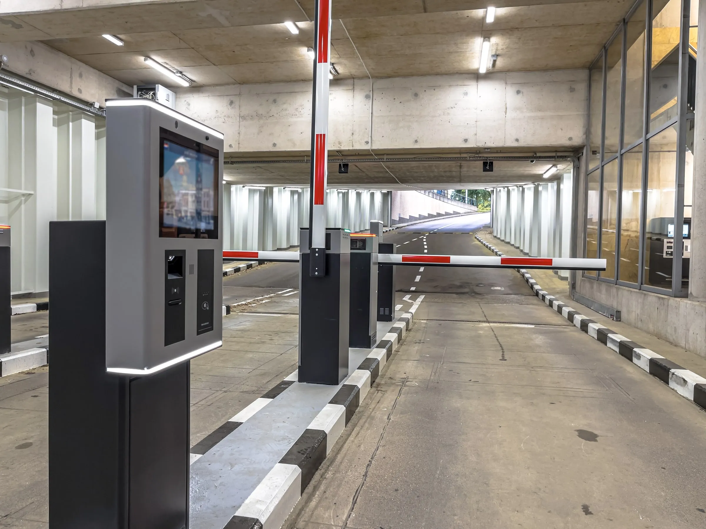 Parking garage entry lane with a pay station kiosk, red and white barrier arms and a ramp rising toward daylight