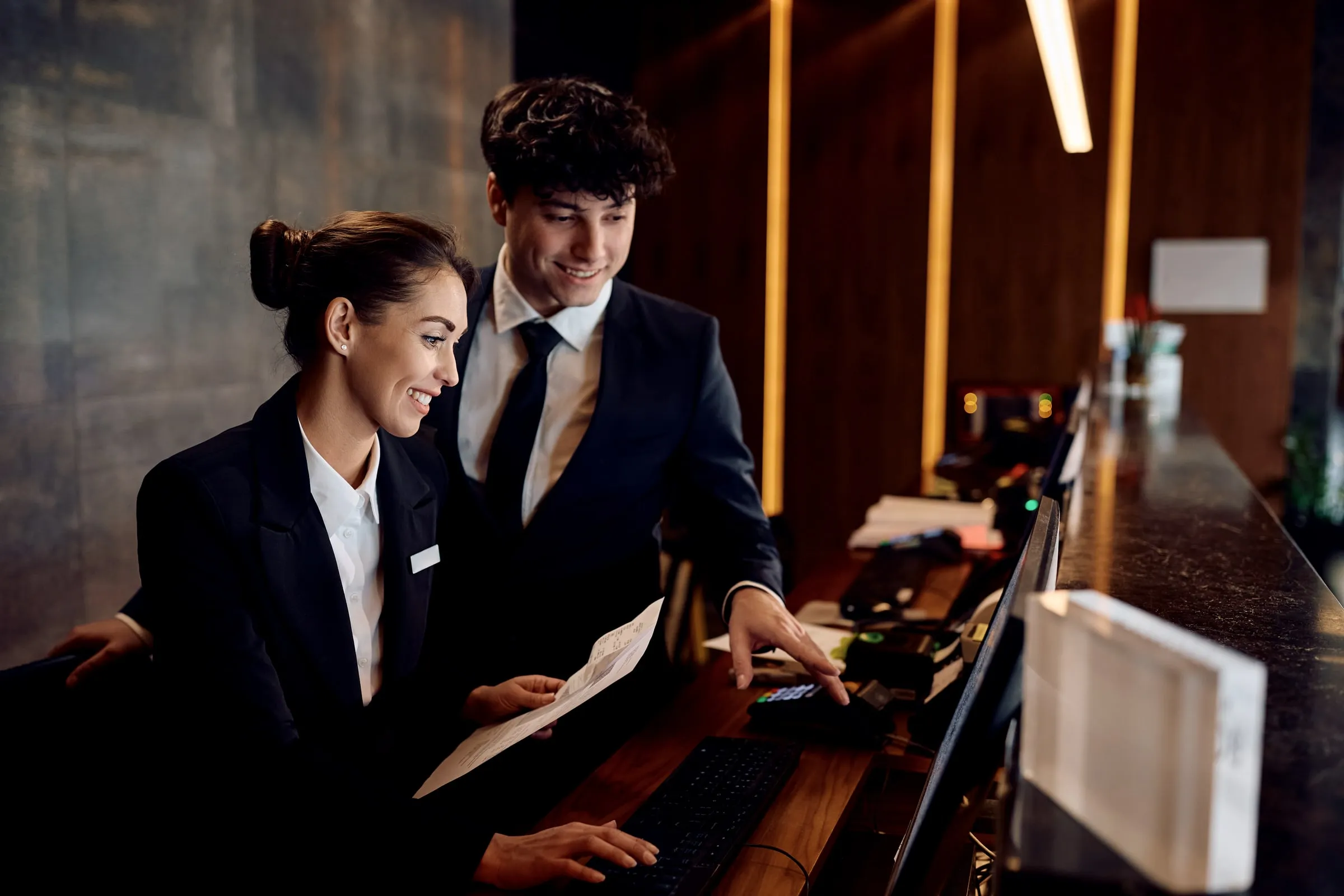 Two front desk agents in dark suits at a hotel reception counter, one seated with a printout and one pointing at a card terminal