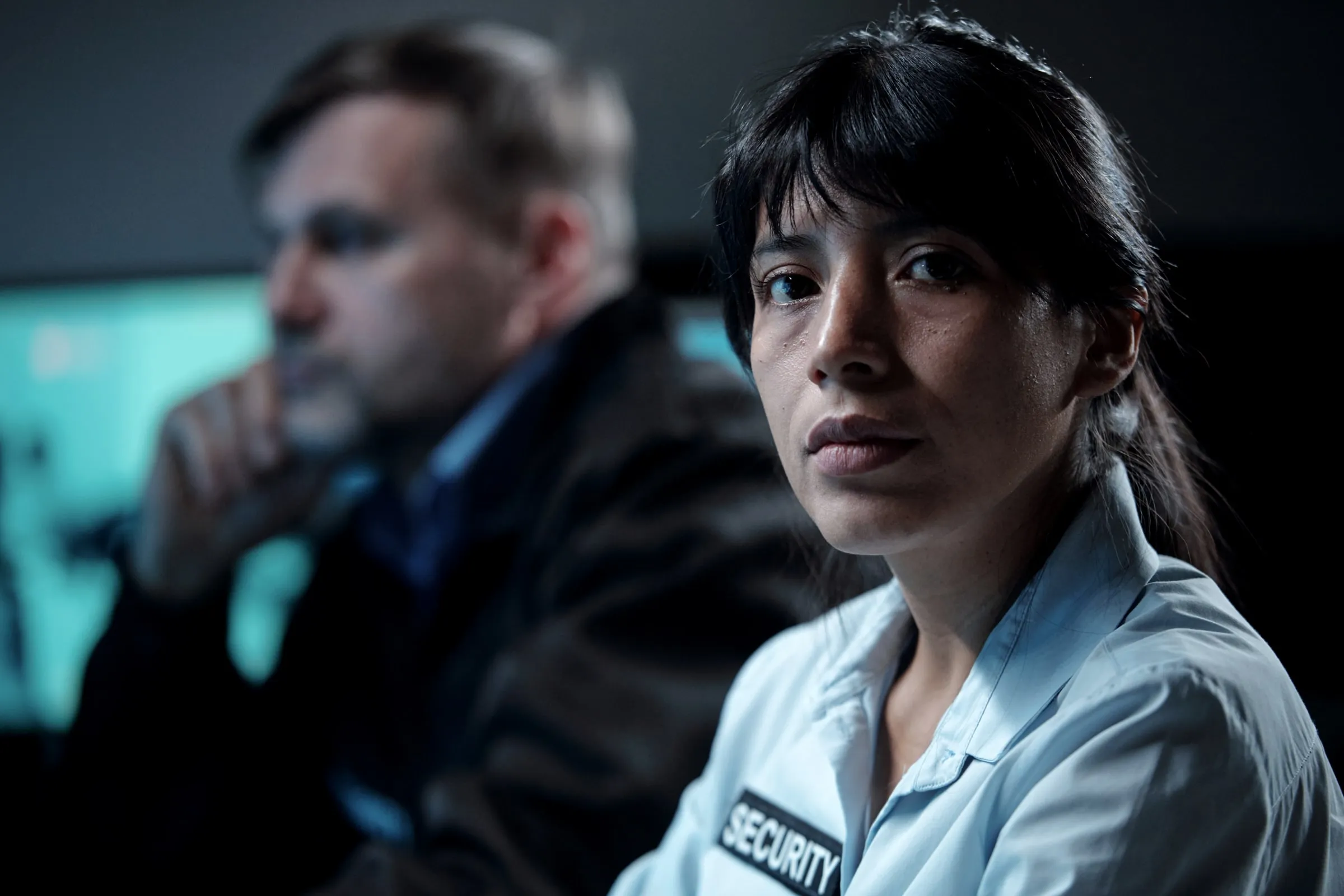 Two security officers in a dim control room, one in a white SECURITY shirt looking at the camera