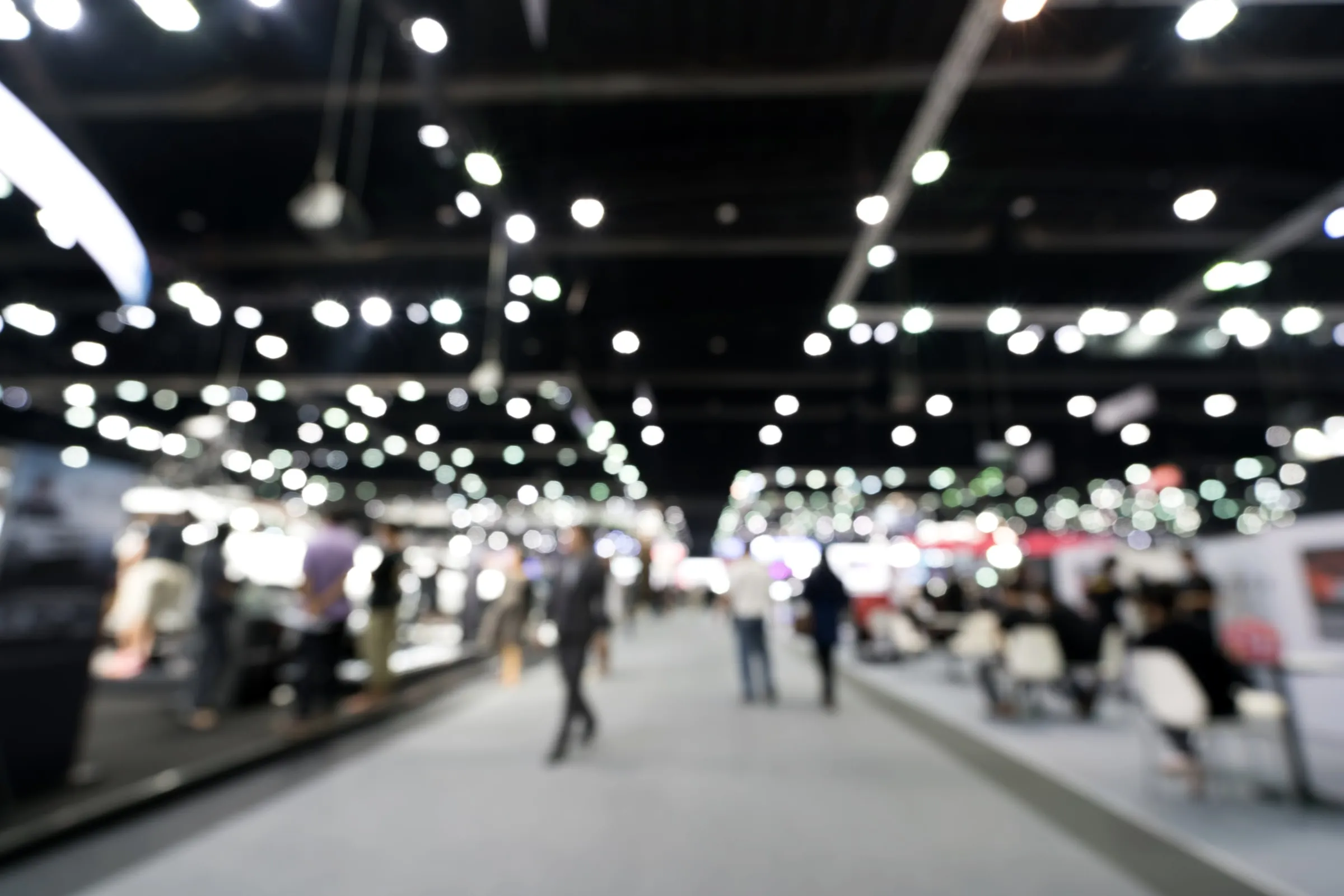 Wide exhibition hall with rows of booths, bright lights and attendees walking the aisles