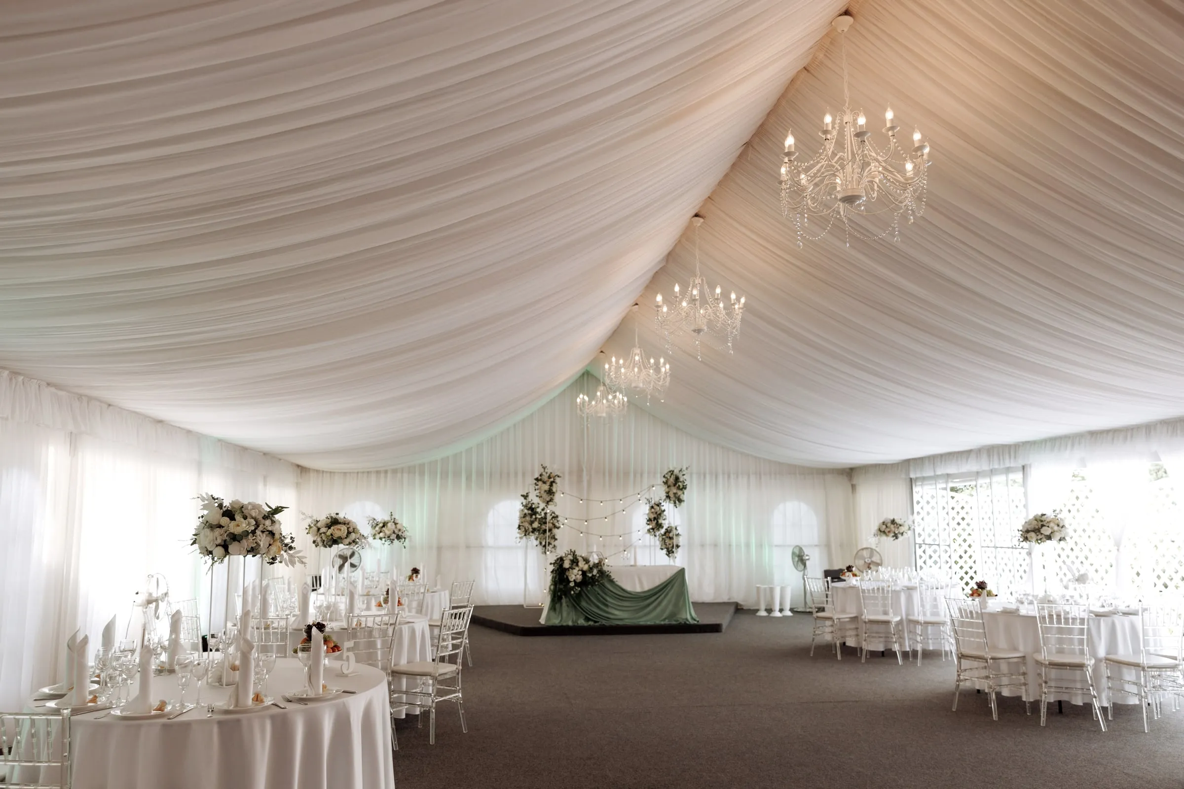Wedding reception tent with draped white ceiling, chandeliers and round tables set for dinner