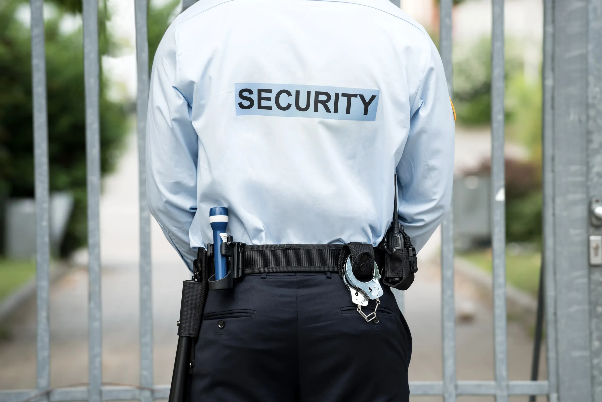 Security officer with SECURITY on the back of a white shirt standing at a metal fence, radio and handcuffs on the belt