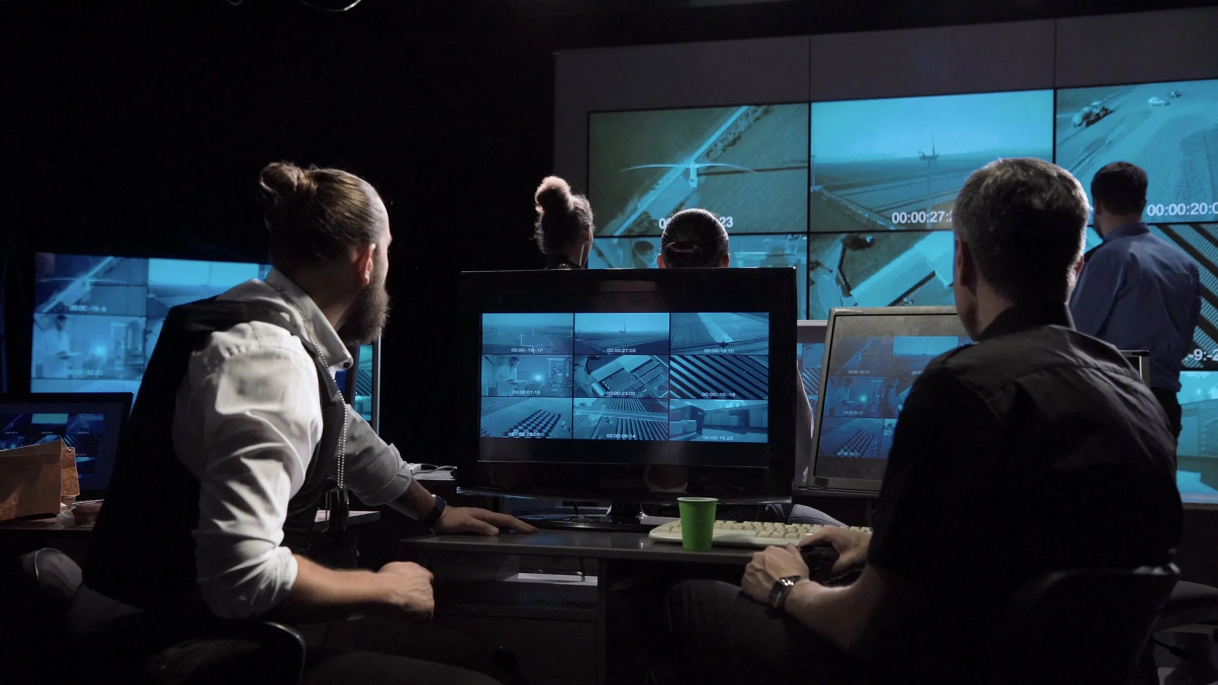 Security team in a monitoring room watching multiple screens showing aerial and interior camera feeds