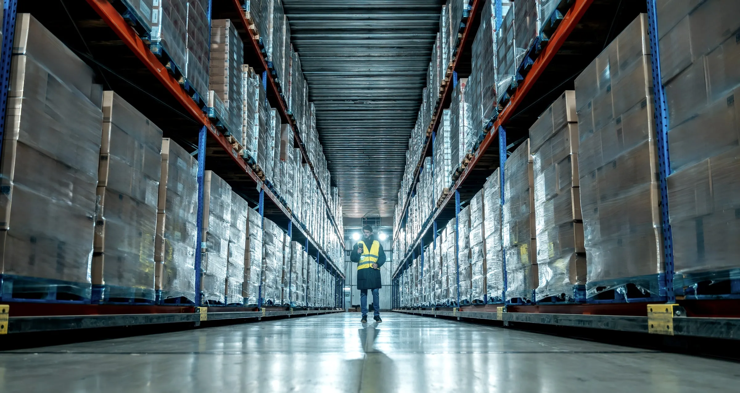 Worker in a hi-vis vest walking a long aisle of a refrigerated warehouse between tall racks of shrink-wrapped pallets