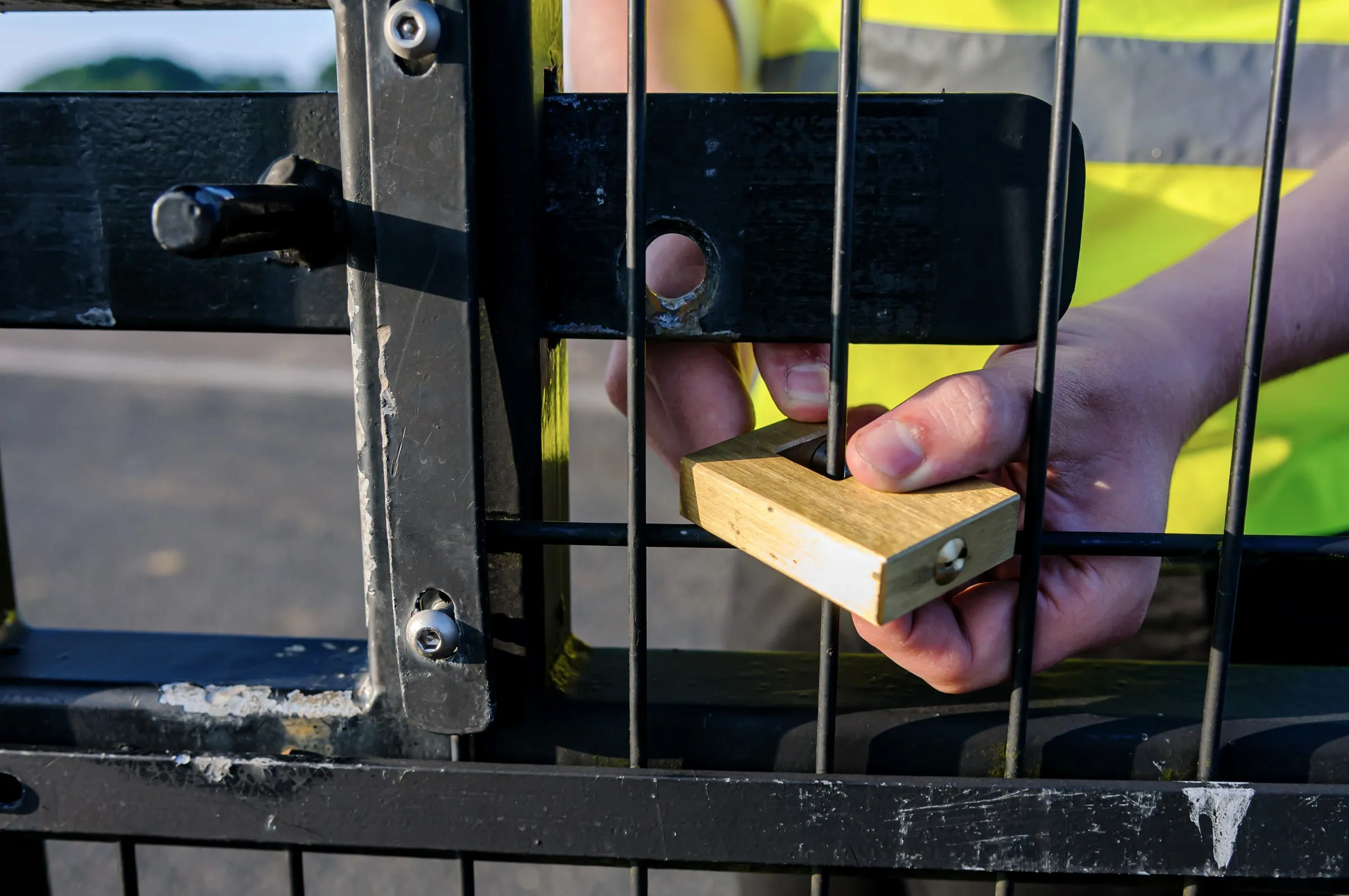 Close-up of a hand fitting a padlock to a chain-link gate, with a person in a yellow hi-vis vest behind it