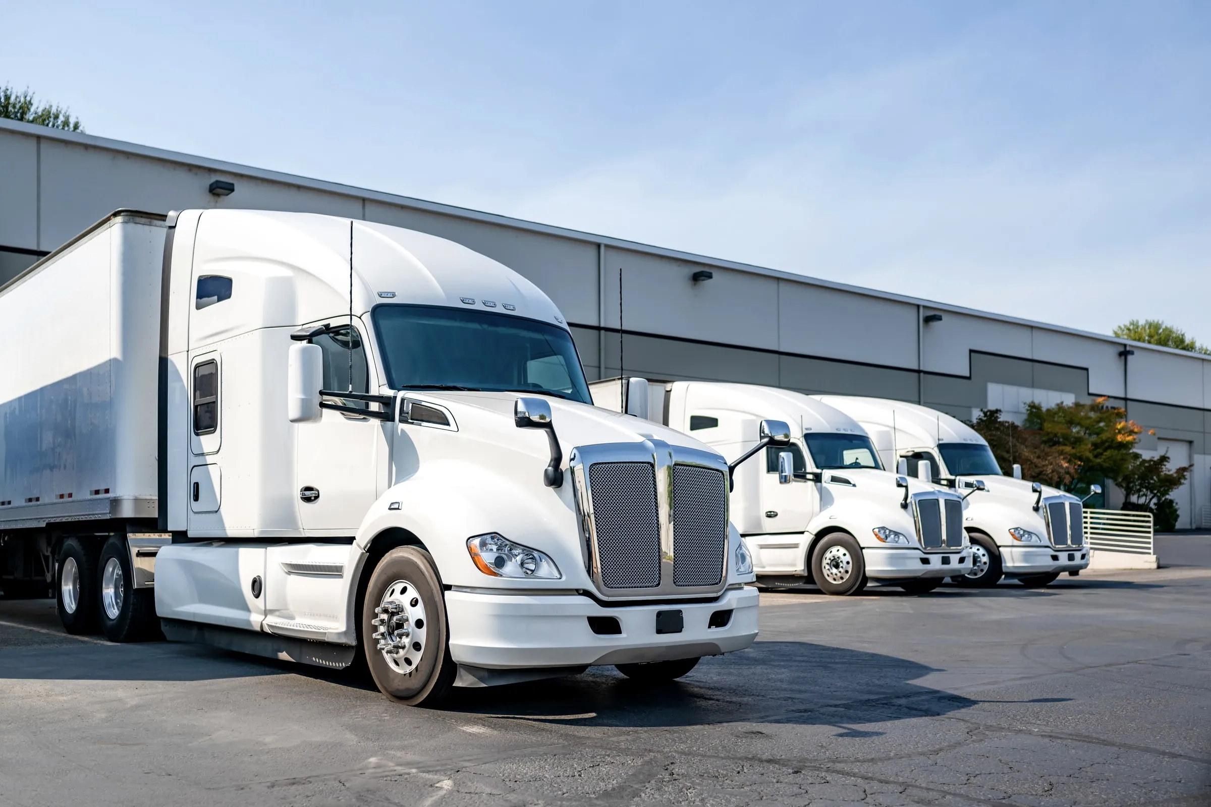 Row of white semi-truck tractors parked nose-out beside a warehouse, with trailers and loading docks behind