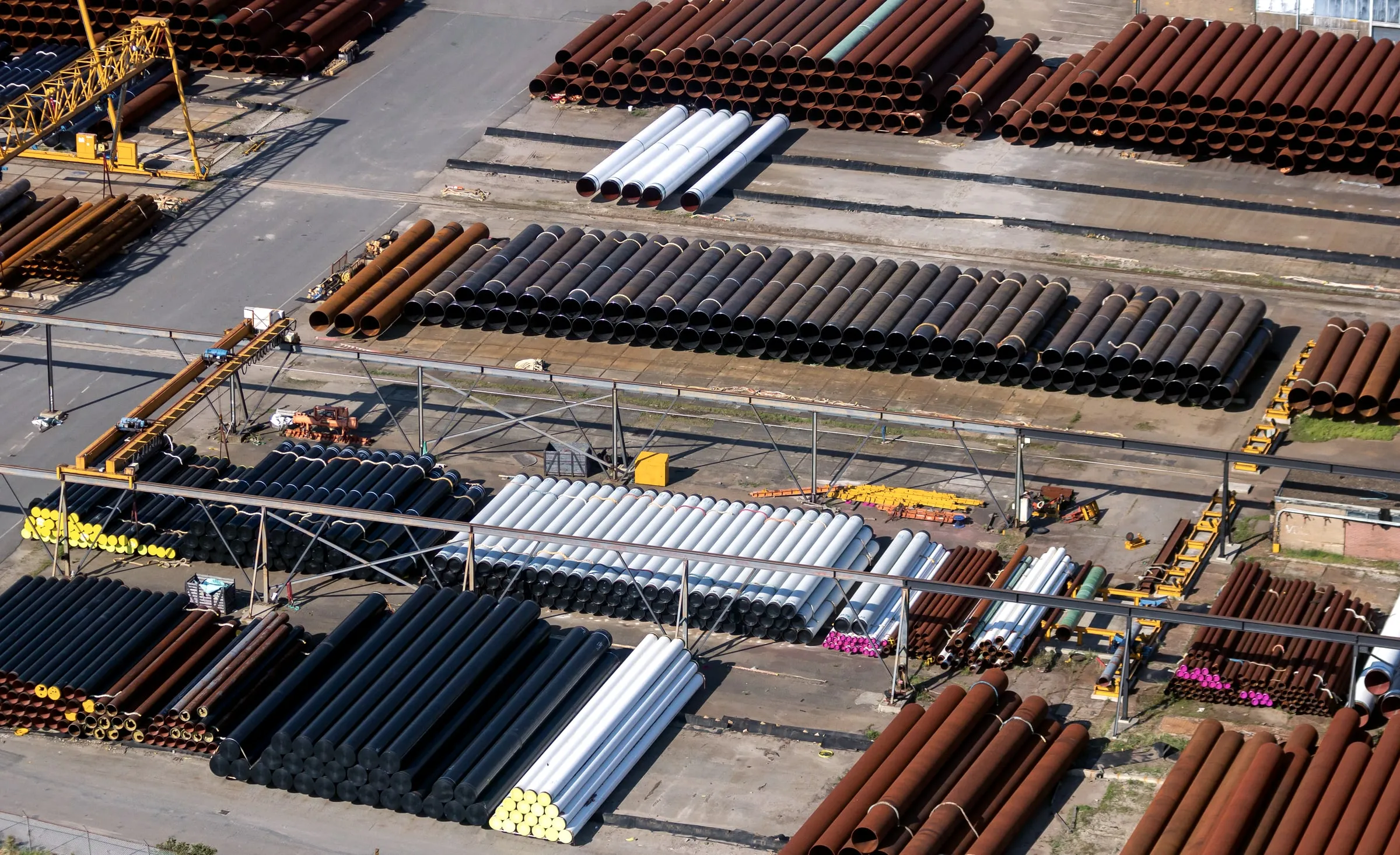 Aerial view of a large pipe laydown yard with long rows of steel pipe sorted by color and a crane at one end
