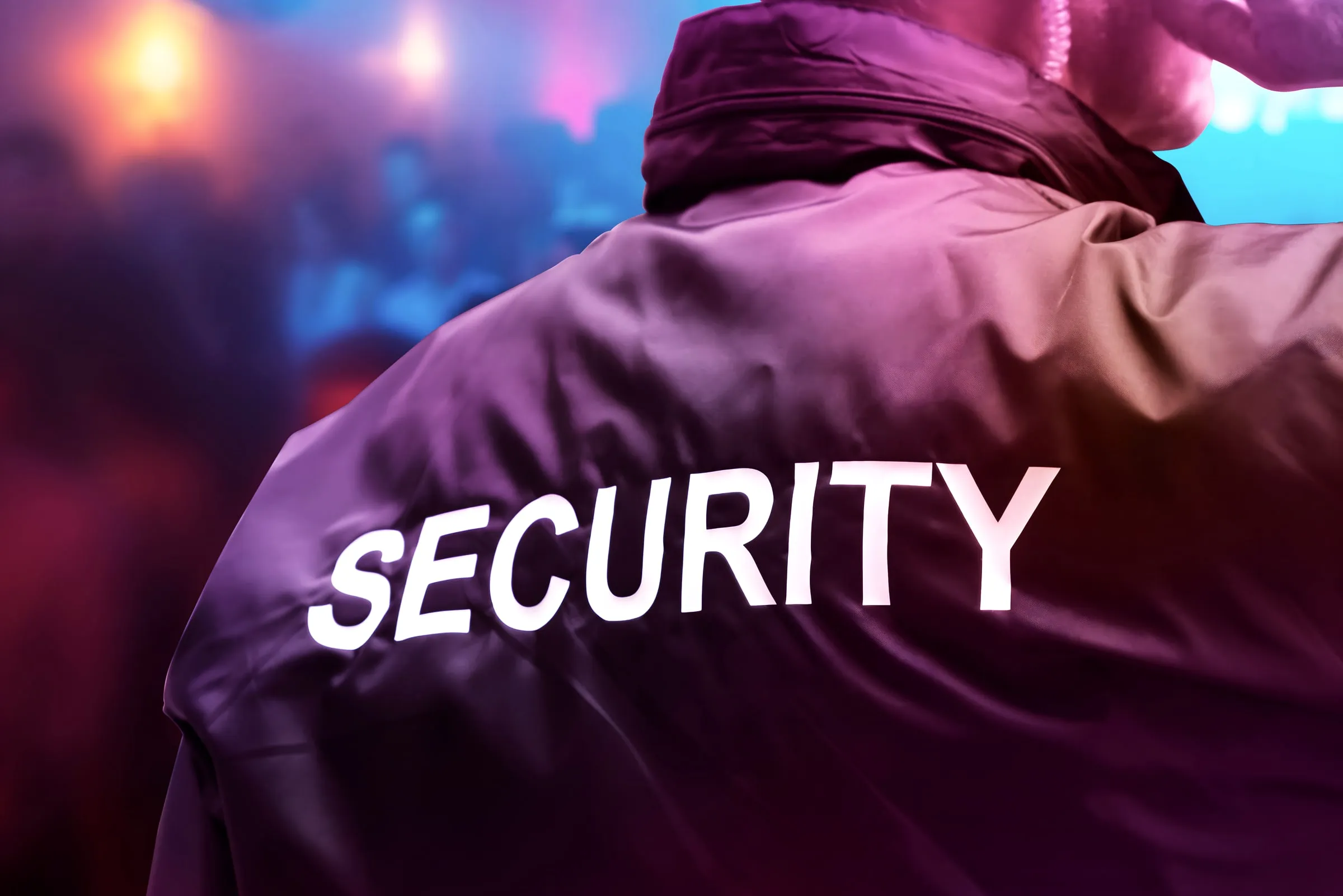 Person wearing a jacket printed Security across the back, seen from behind against a blurred colored background