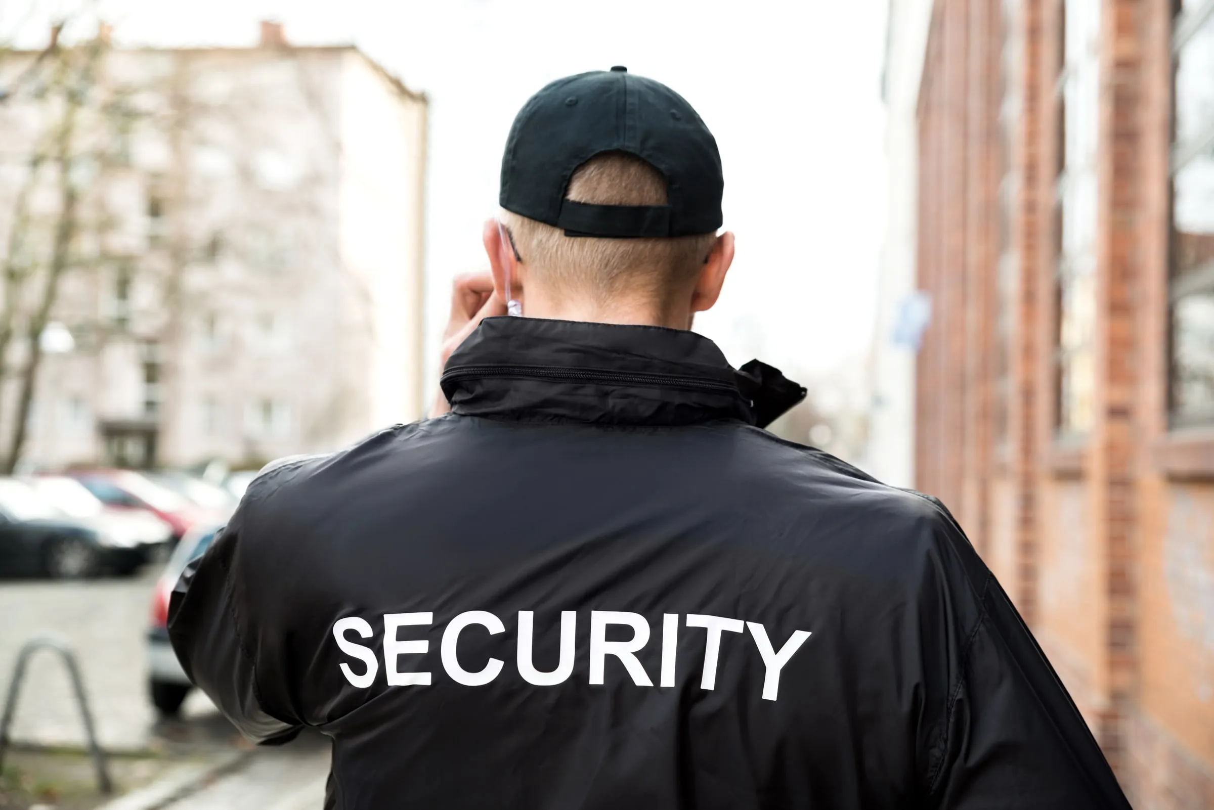 Security officer seen from behind wearing a cap and jacket printed Security, standing on an urban sidewalk