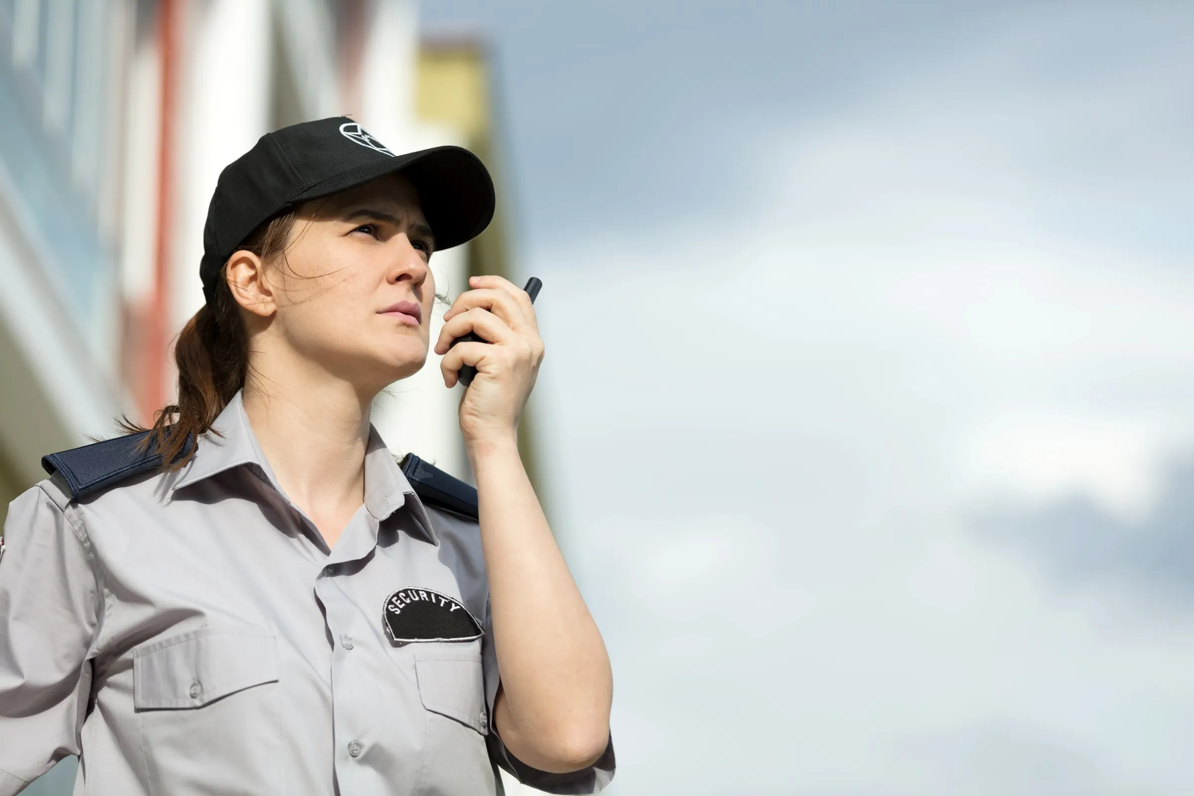 Female security officer wearing a cap and radio, speaking into a handheld radio outside a building