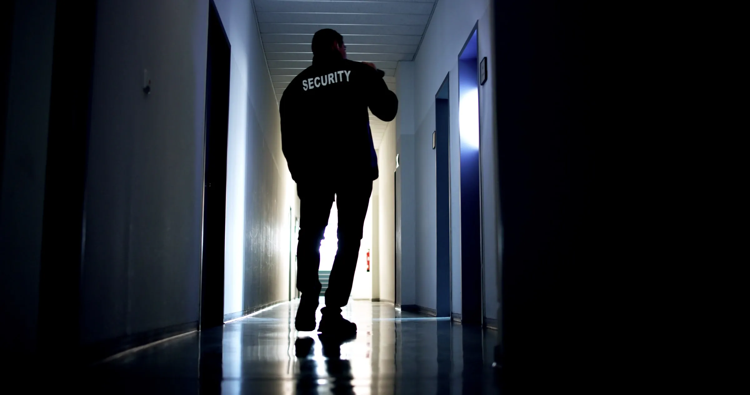Silhouette of a security officer wearing a jacket printed Security, walking down a lit interior corridor