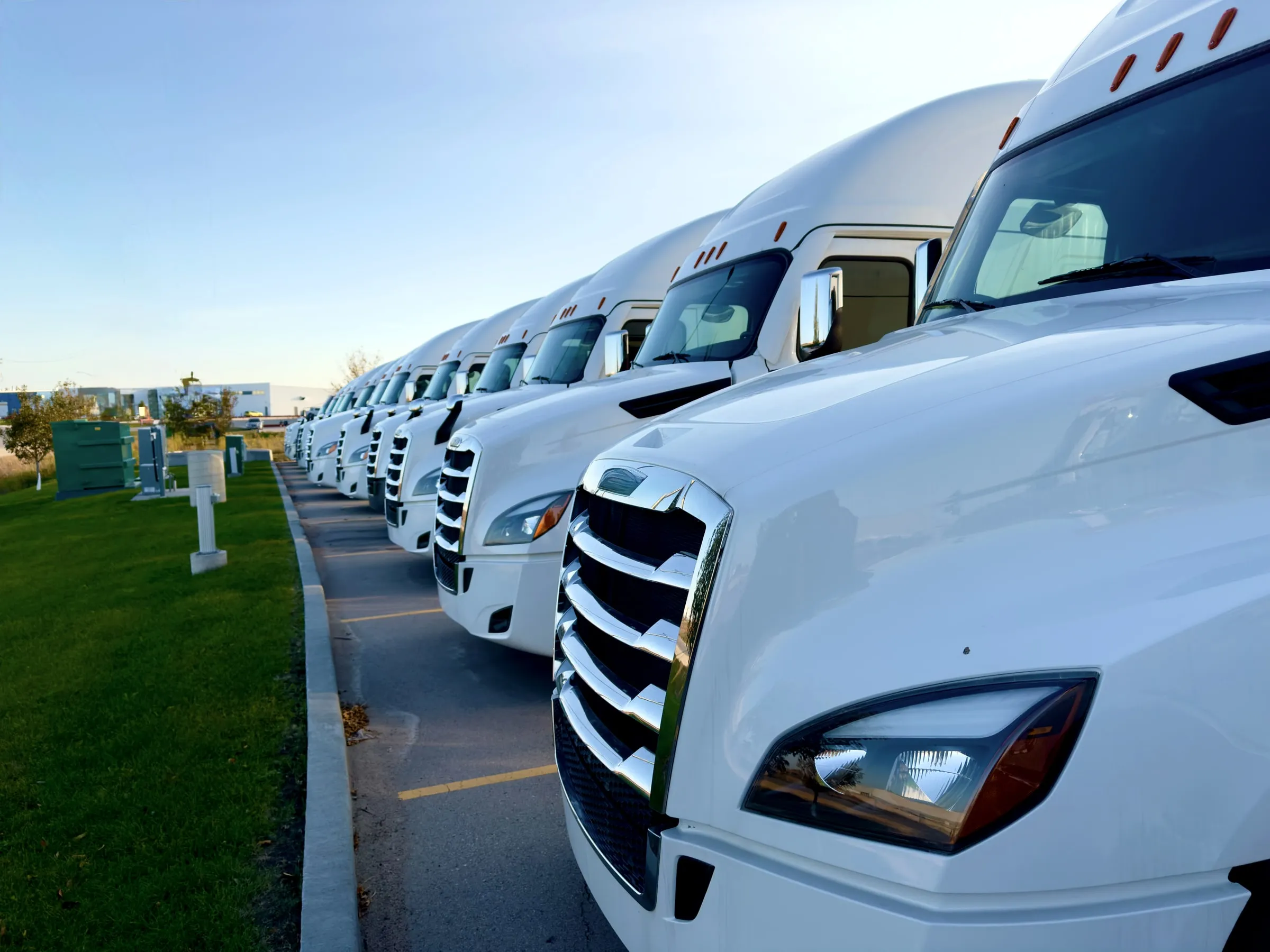 Row of white semi-truck cabs parked front-facing in a company lot beside a grass strip and low buildings