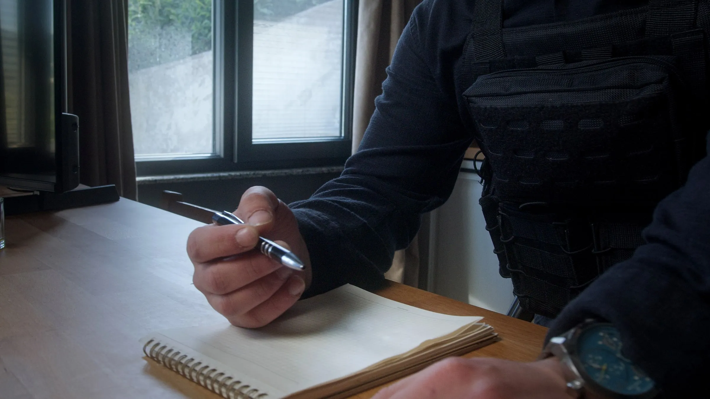 Close-up of an officer's hand writing in a notebook at a desk, lit by a small lamp