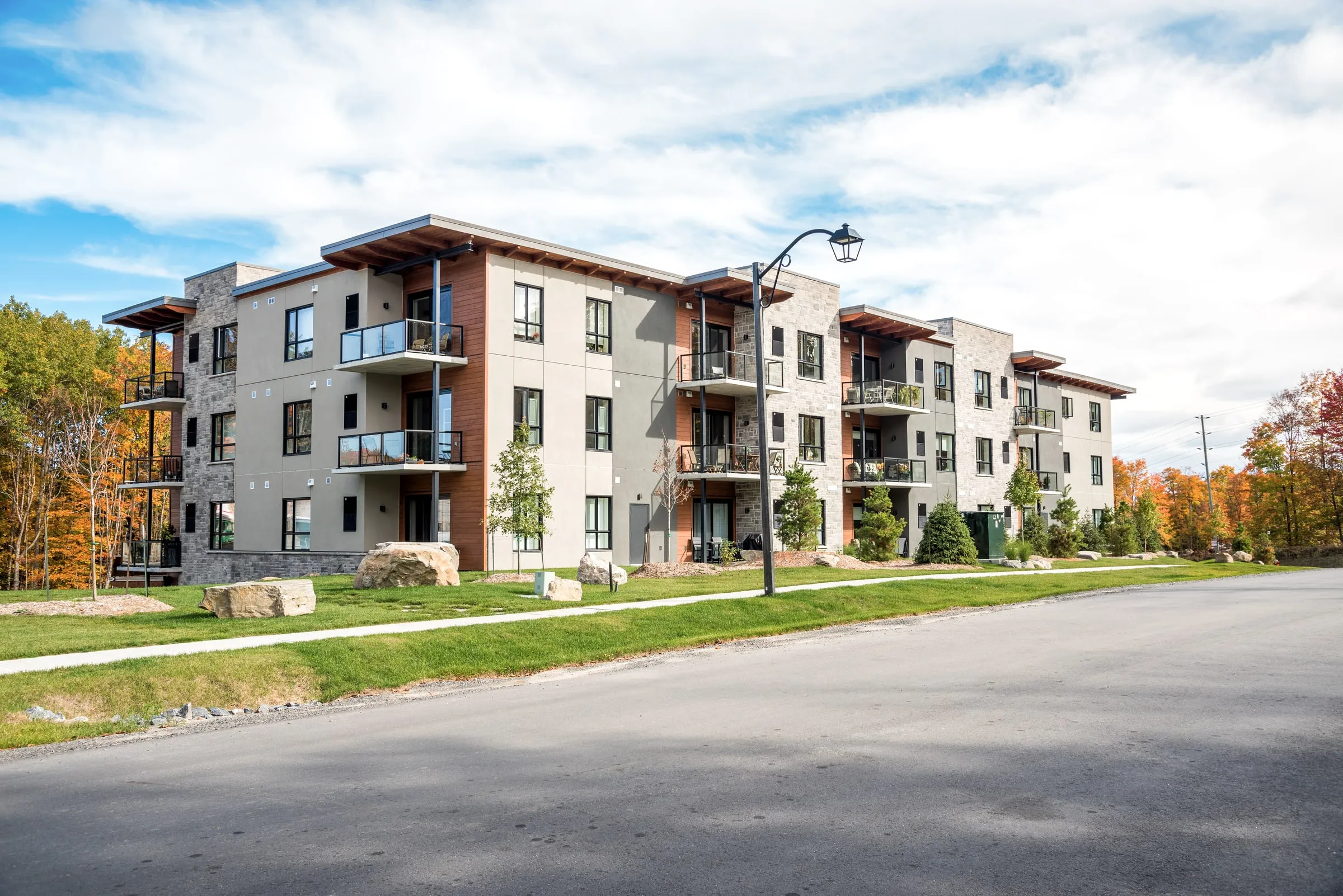 Three-storey apartment building with grey stucco, stone and wood-look siding, glass balconies and new landscaping with boulders along a quiet street in autumn