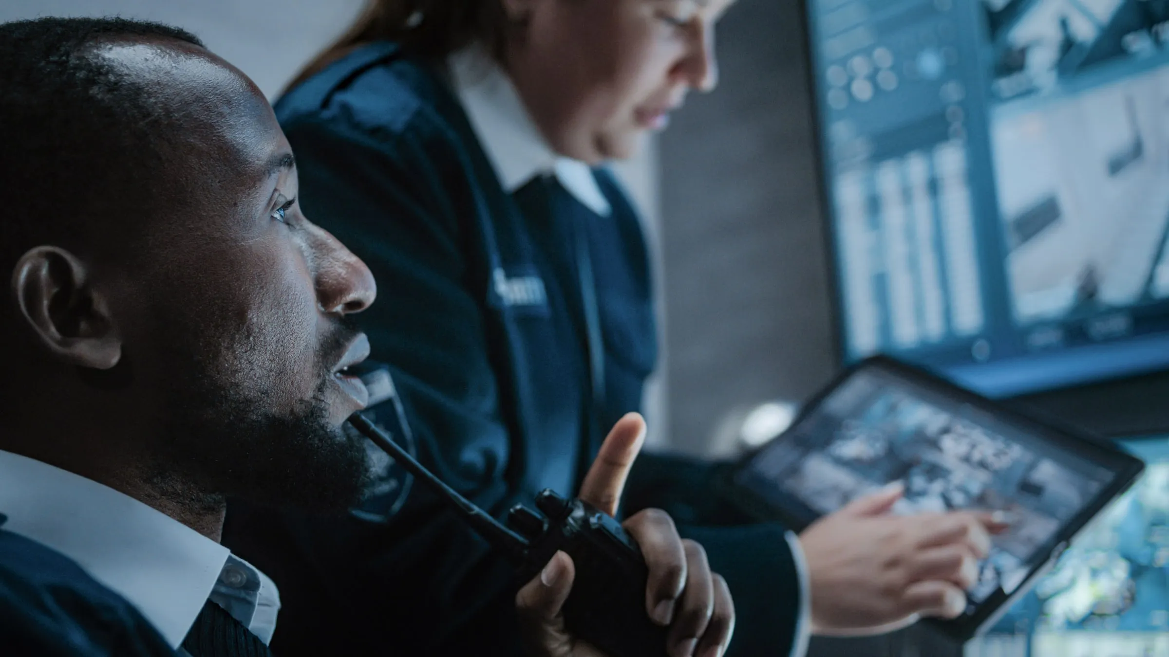 Two officers in dark uniforms in a control room, one speaking into a handheld radio while the other studies a tablet in front of camera monitors