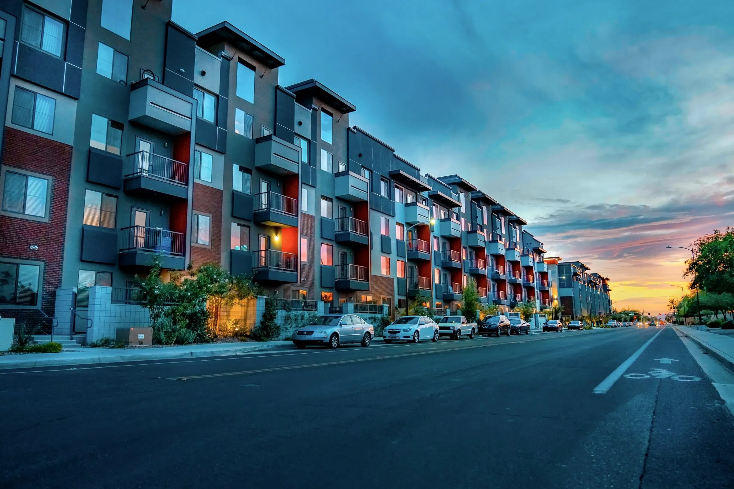 Modern five-storey apartment block with grey and red panels and balconies at dusk, with cars parked along the street under a sunset sky