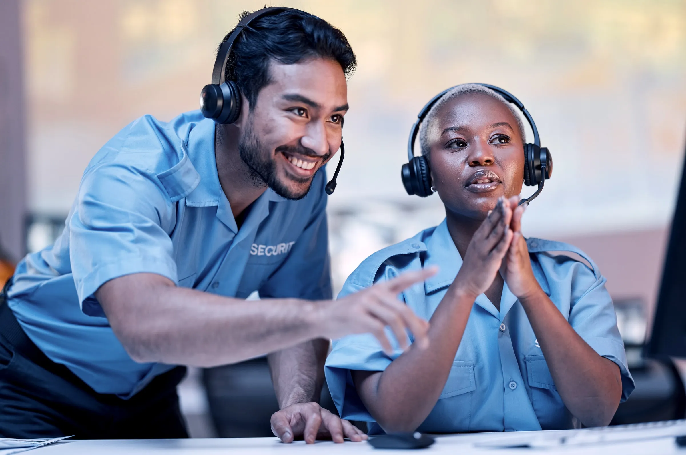 Two officers in light blue SECURITY shirts and headsets at a desk, one standing and pointing at a screen while the other listens with hands clasped
