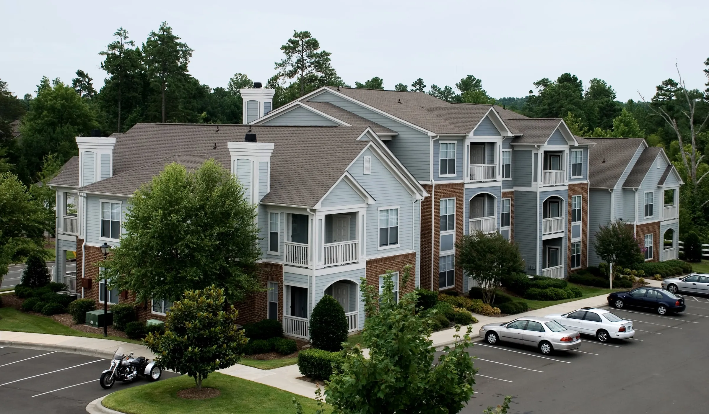 Elevated view of a three-storey blue-grey and brick apartment building with white balconies beside a surface parking lot with parked cars and a motorcycle