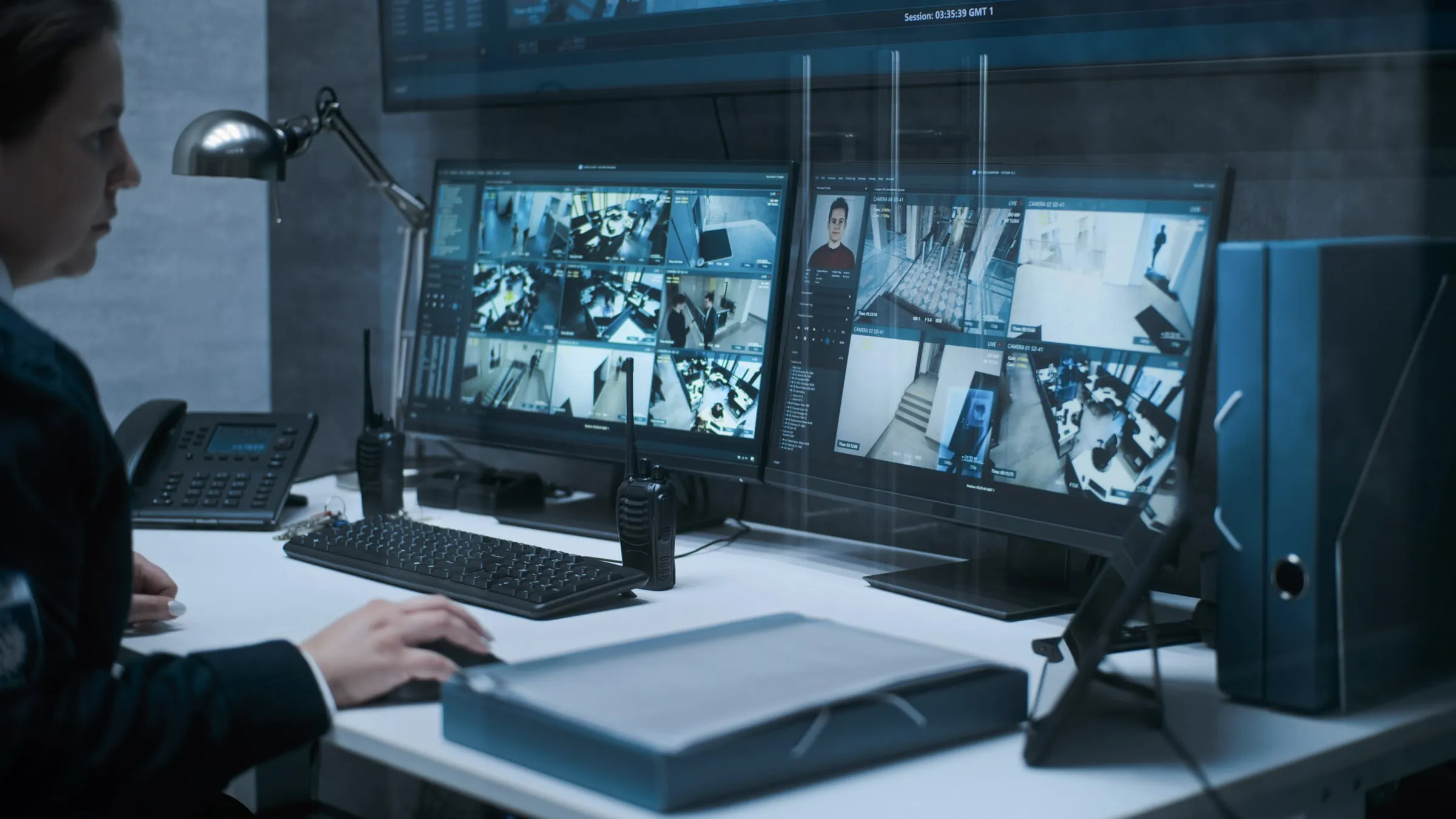Officer seated at a desk with two monitors showing grids of camera feeds, a wall screen above, two radios and a desk phone in a dim room