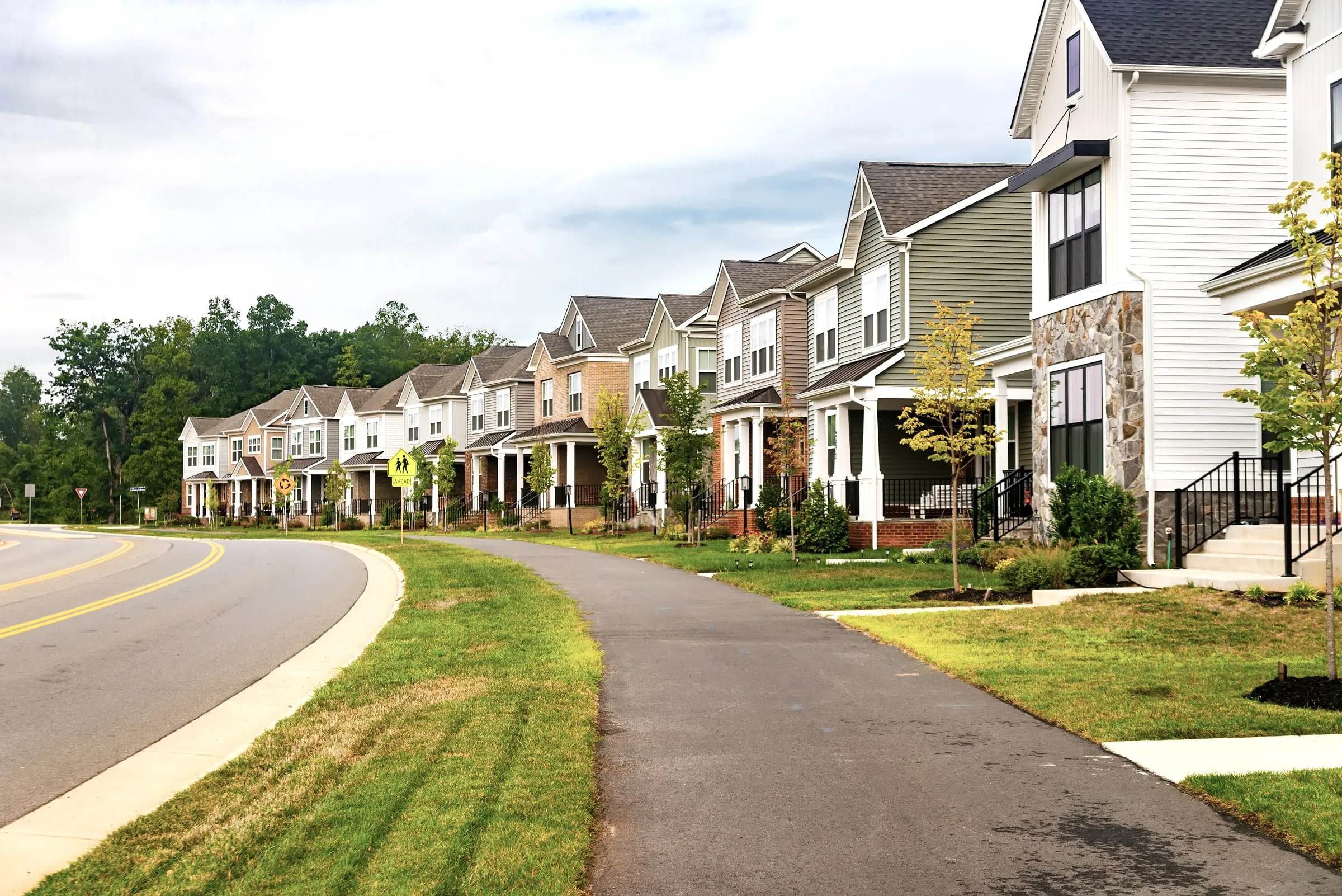 Row of new two-storey houses with front porches and mixed siding colors along a curving street with a sidewalk, young trees and a school crossing sign