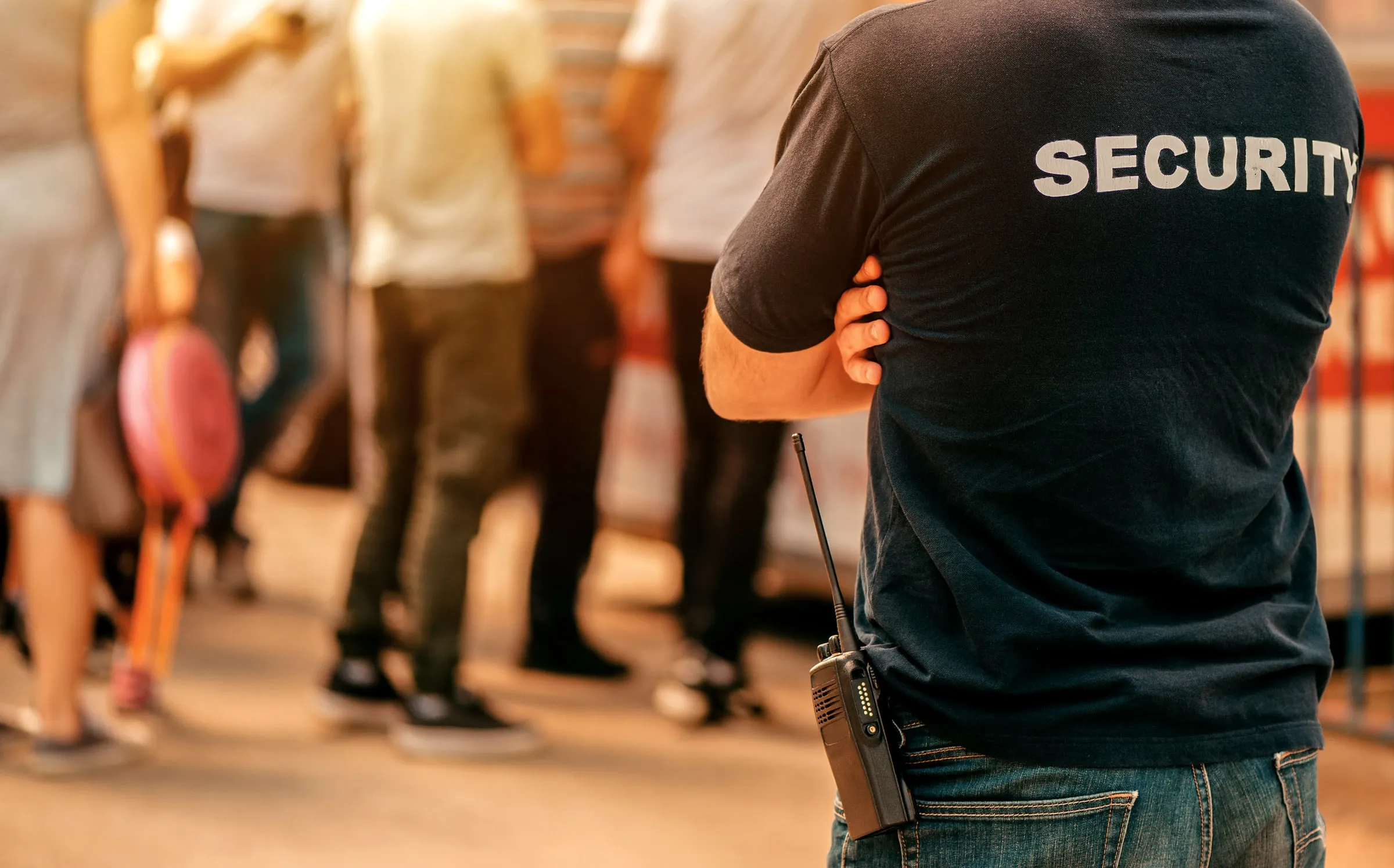 Back of a security officer in a black SECURITY shirt with a radio, in a crowded space
