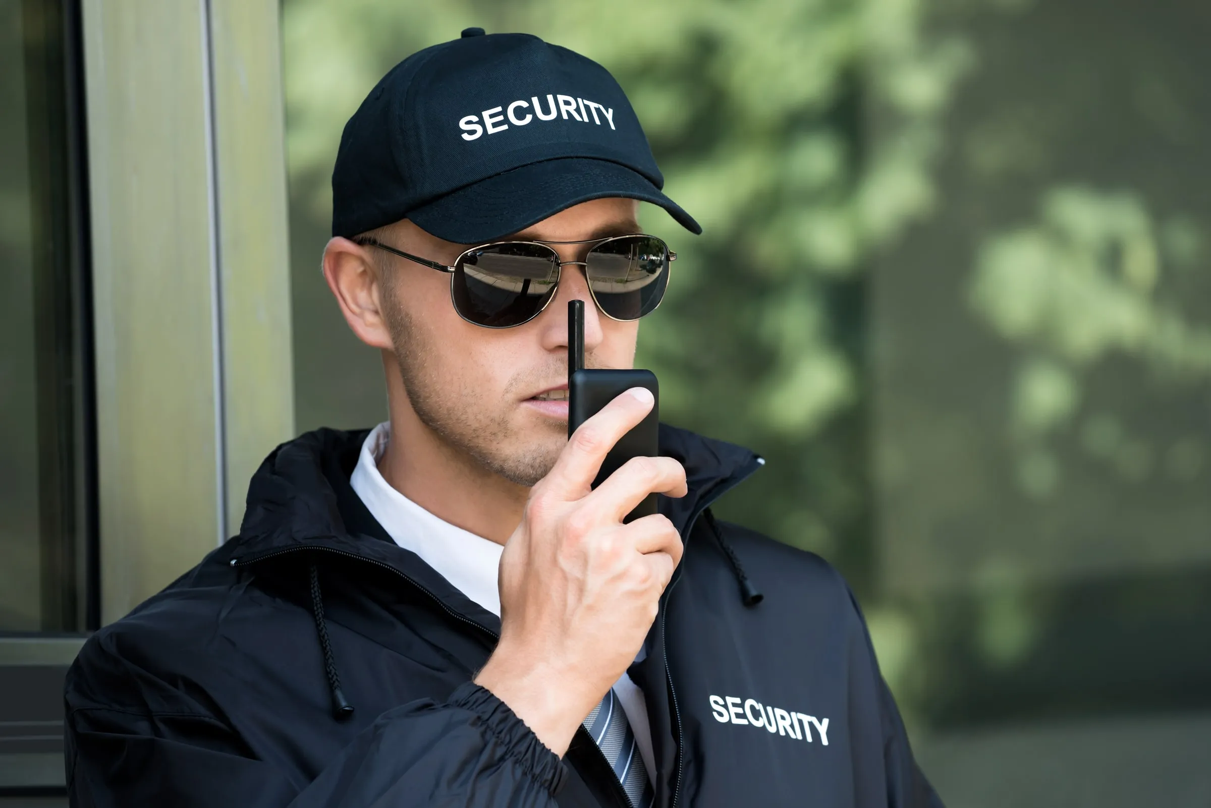 Security officer in a black SECURITY cap and jacket speaking into a handheld radio outside a glass entrance