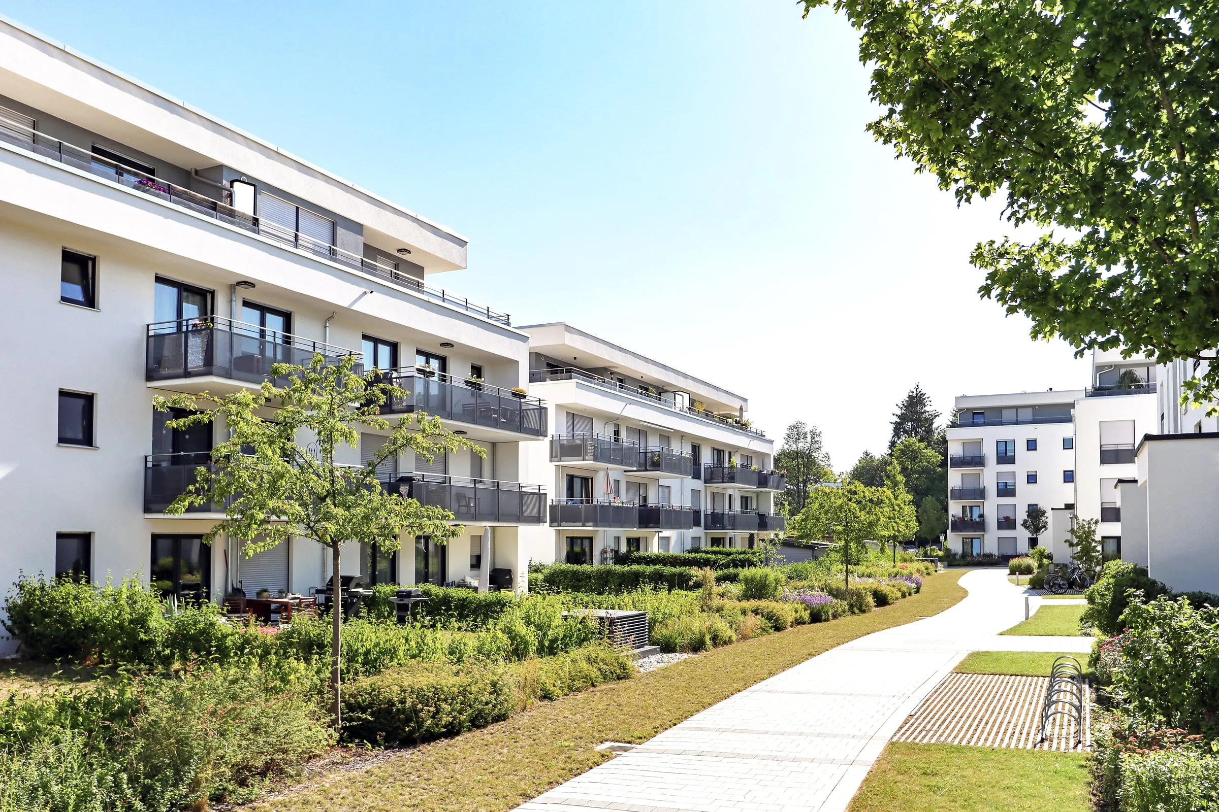 Low-rise apartment buildings with balconies and landscaped courtyards along a paved path on a sunny afternoon