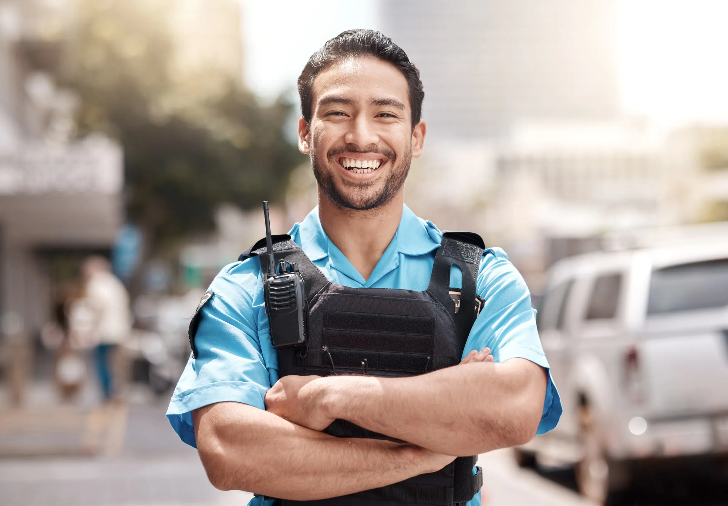 Smiling security officer in a light blue shirt and black tactical vest with a radio, arms folded, on a city street beside a parked truck