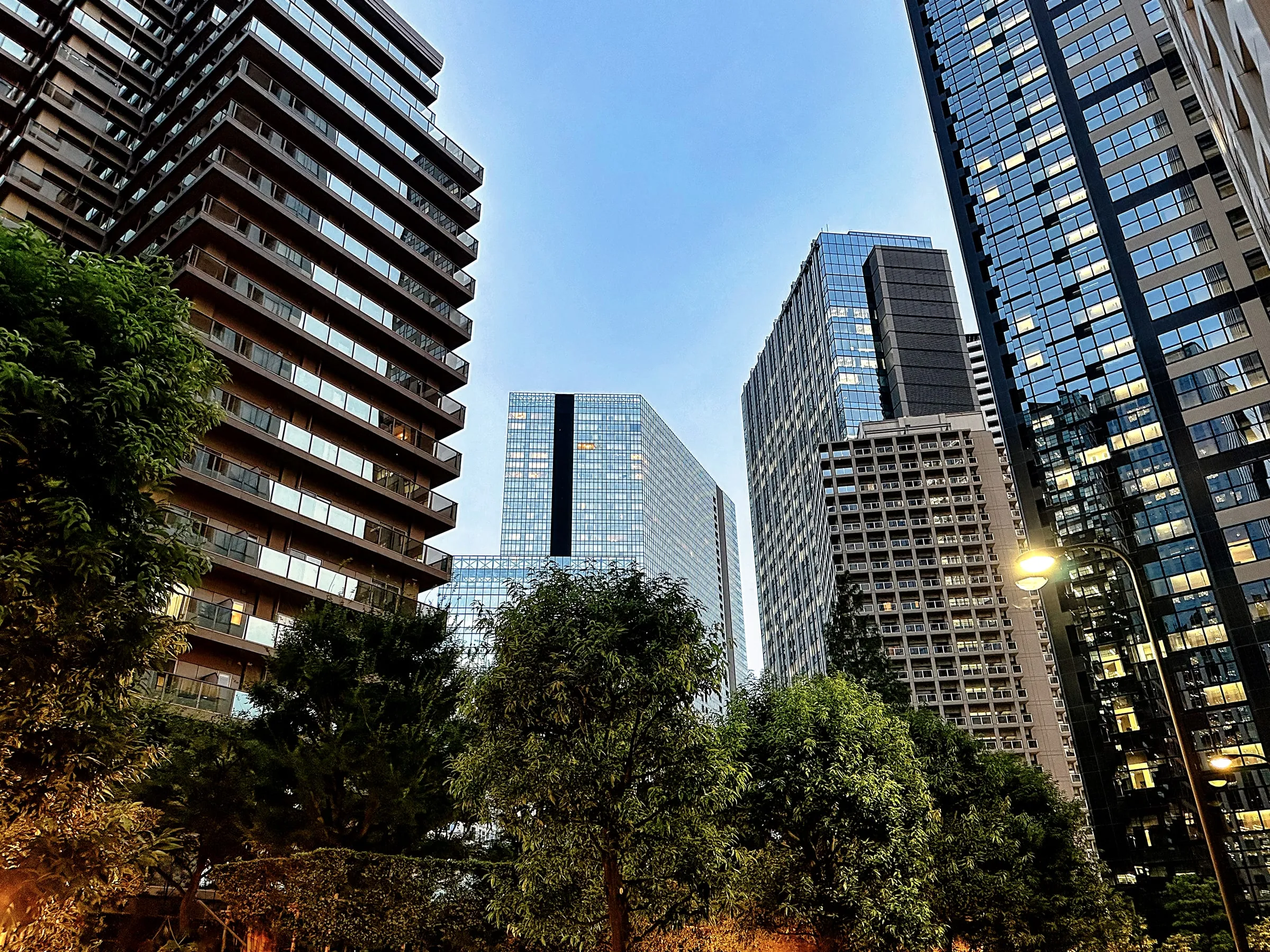 Glass residential towers with stacked balconies rising above a tree-lined courtyard at dusk, with windows lit and a street lamp glowing