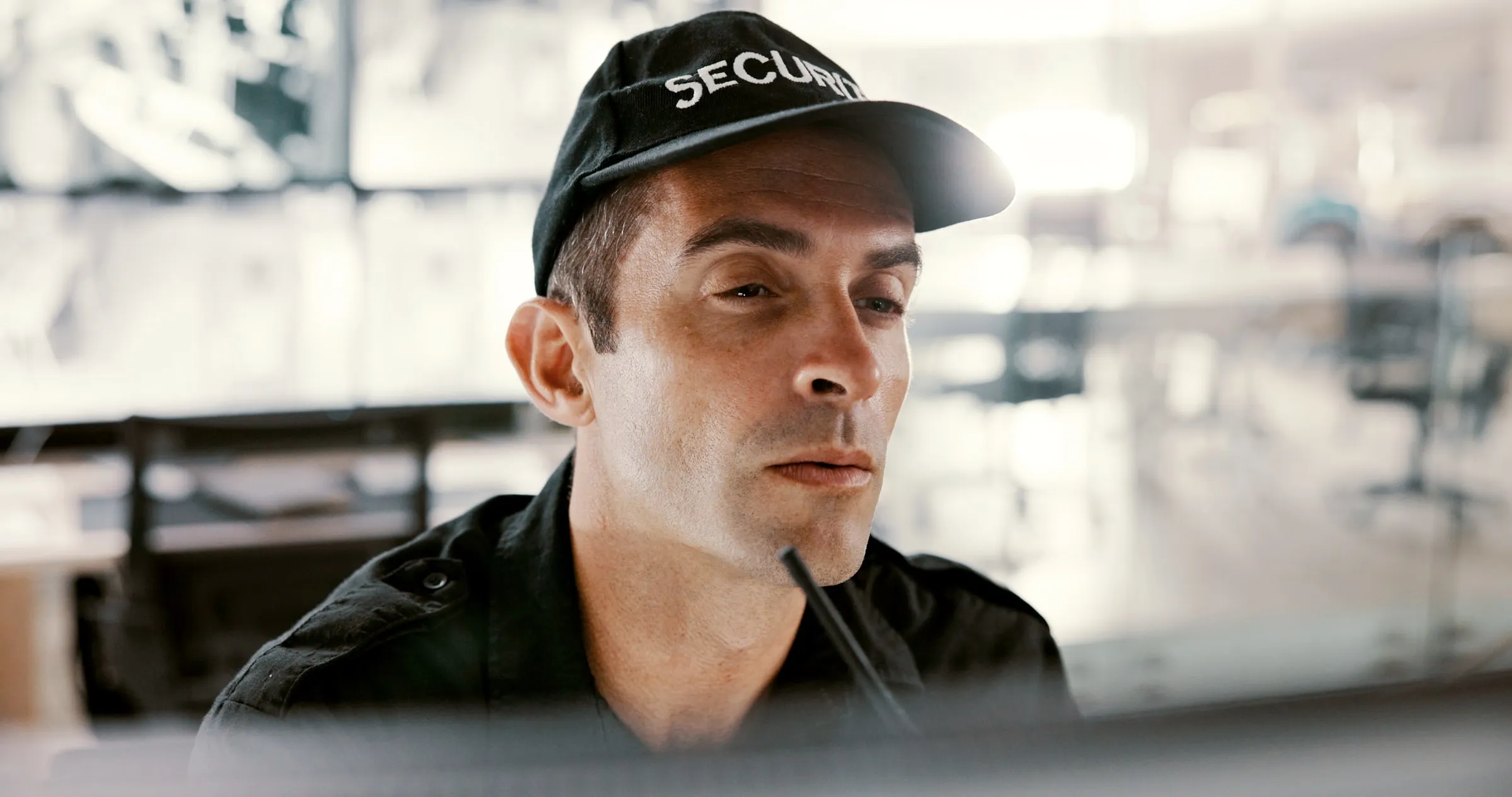 Close view of a security officer in a black SECURITY cap and shirt looking intently at a screen, with a bright office blurred behind him
