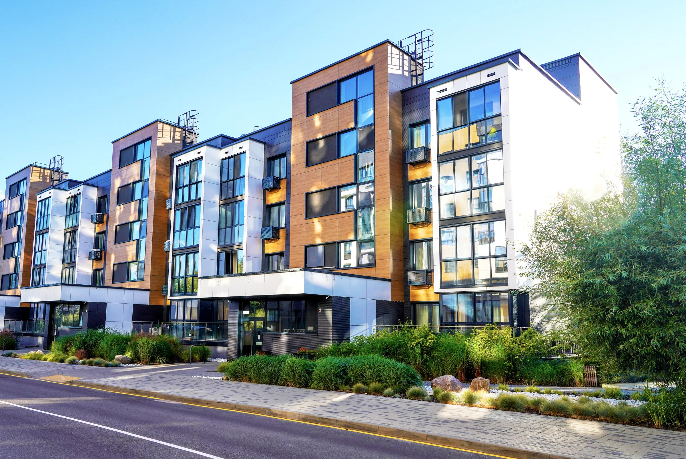 New four-storey apartment building with wood-look and white panels, large windows and fresh landscaping of ornamental grasses and boulders beside a quiet road