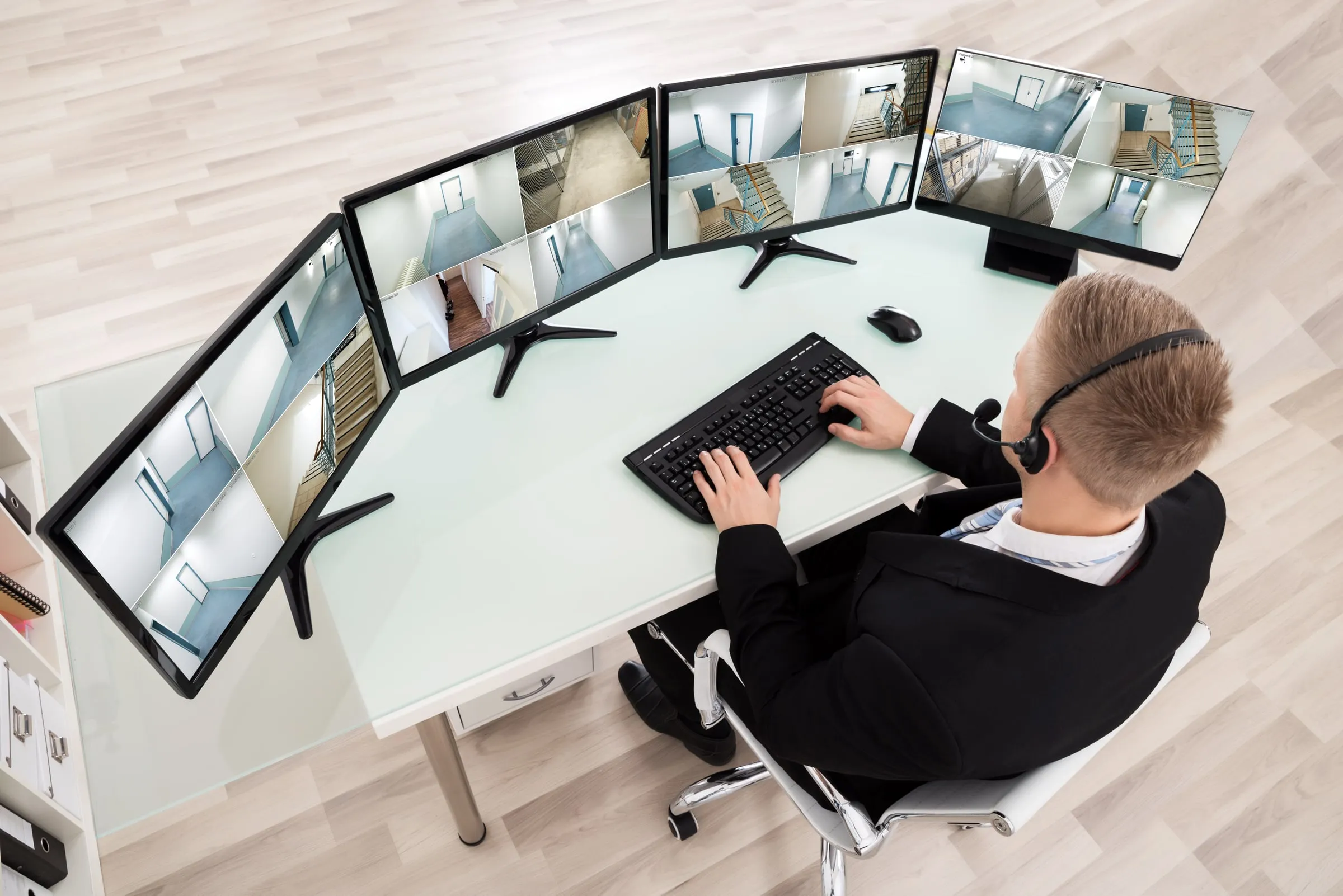 Man in a dark suit and headset at a curved white desk watching a bank of monitors showing camera views of corridors and stairwells