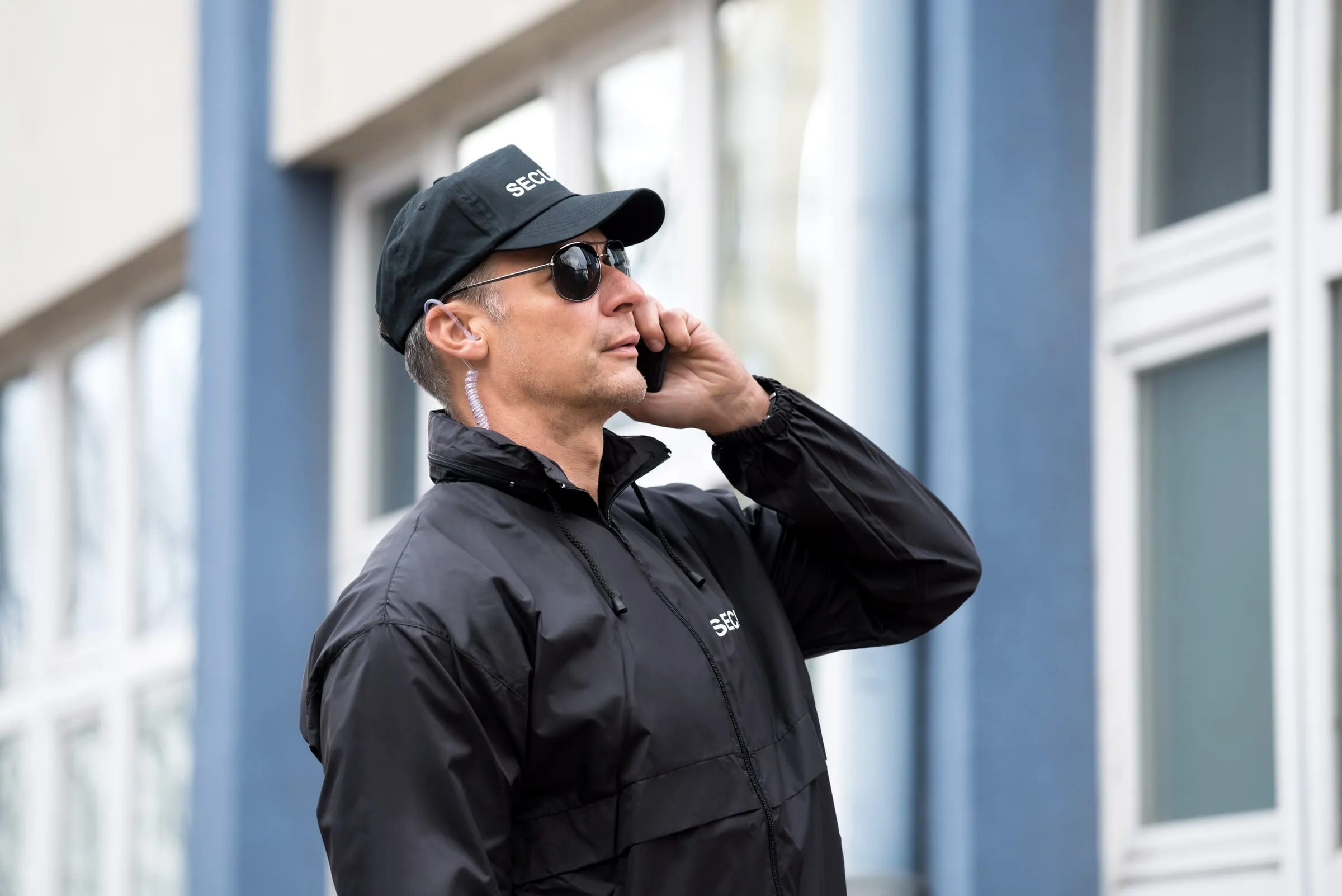 Security officer in a black SECURITY cap, sunglasses and jacket with an earpiece, talking on a mobile phone outside a building with blue columns