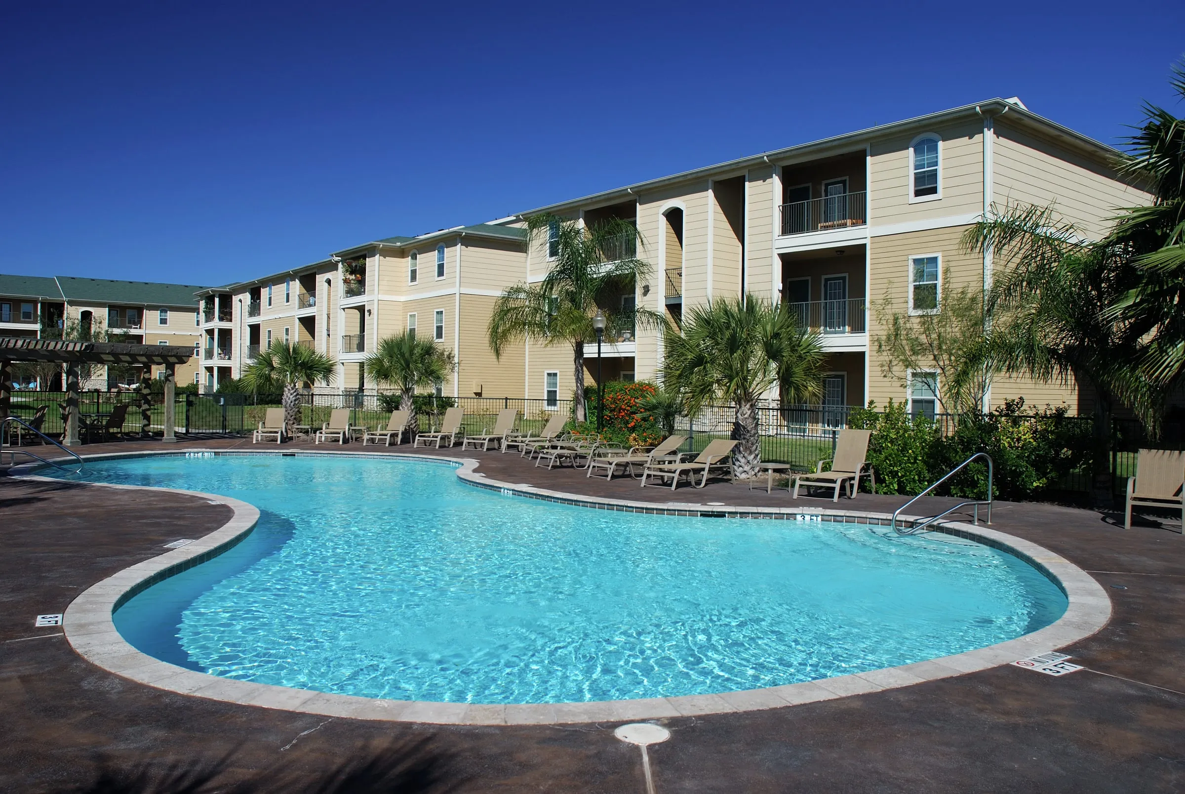 Curved outdoor swimming pool with clear blue water, a row of lounge chairs, palm trees and a black iron fence in front of tan three-storey apartment buildings under a deep blue sky
