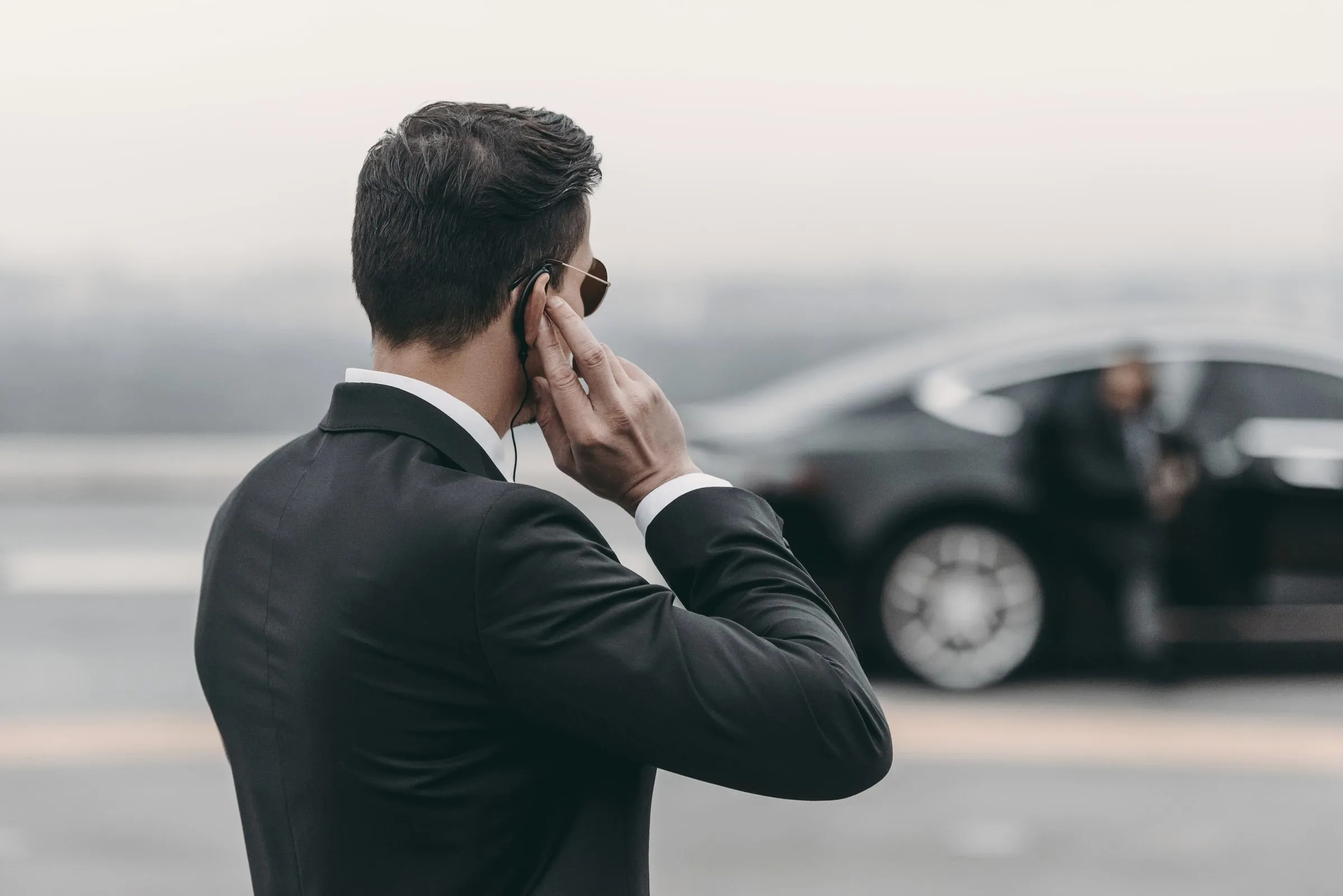 Officer in a dark suit and sunglasses touching an earpiece while a black sedan waits behind him