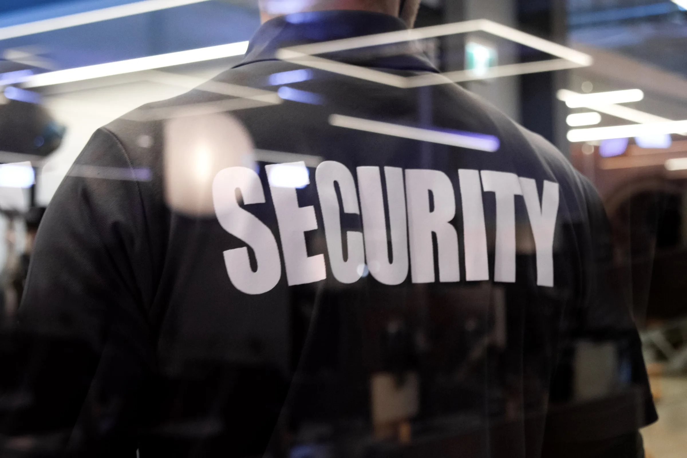 Back of a security officer in a dark SECURITY shirt seen through a glass door with reflected ceiling lights