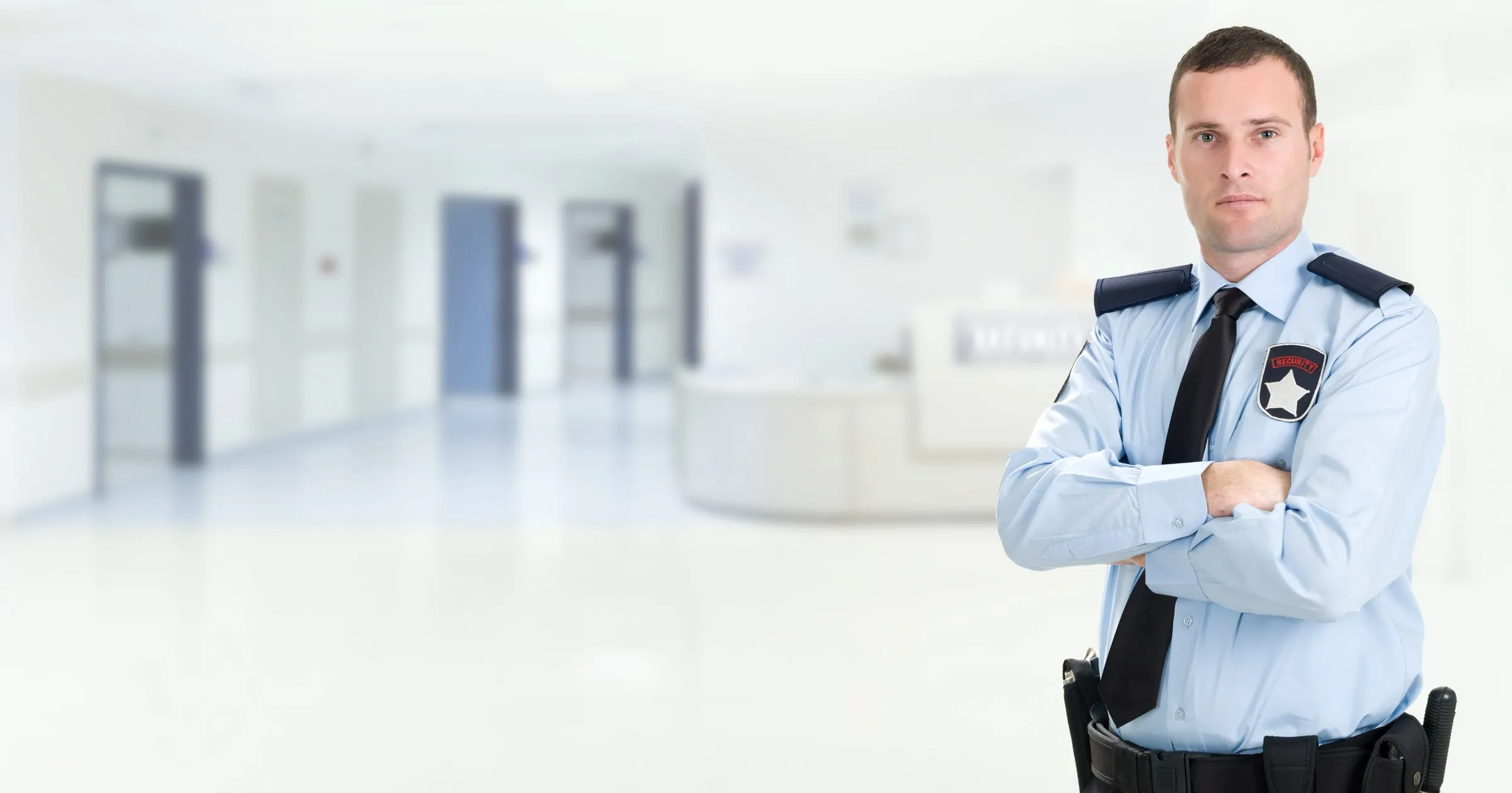 Security officer in a light blue uniform shirt with a badge, standing in a bright hallway with arms crossed