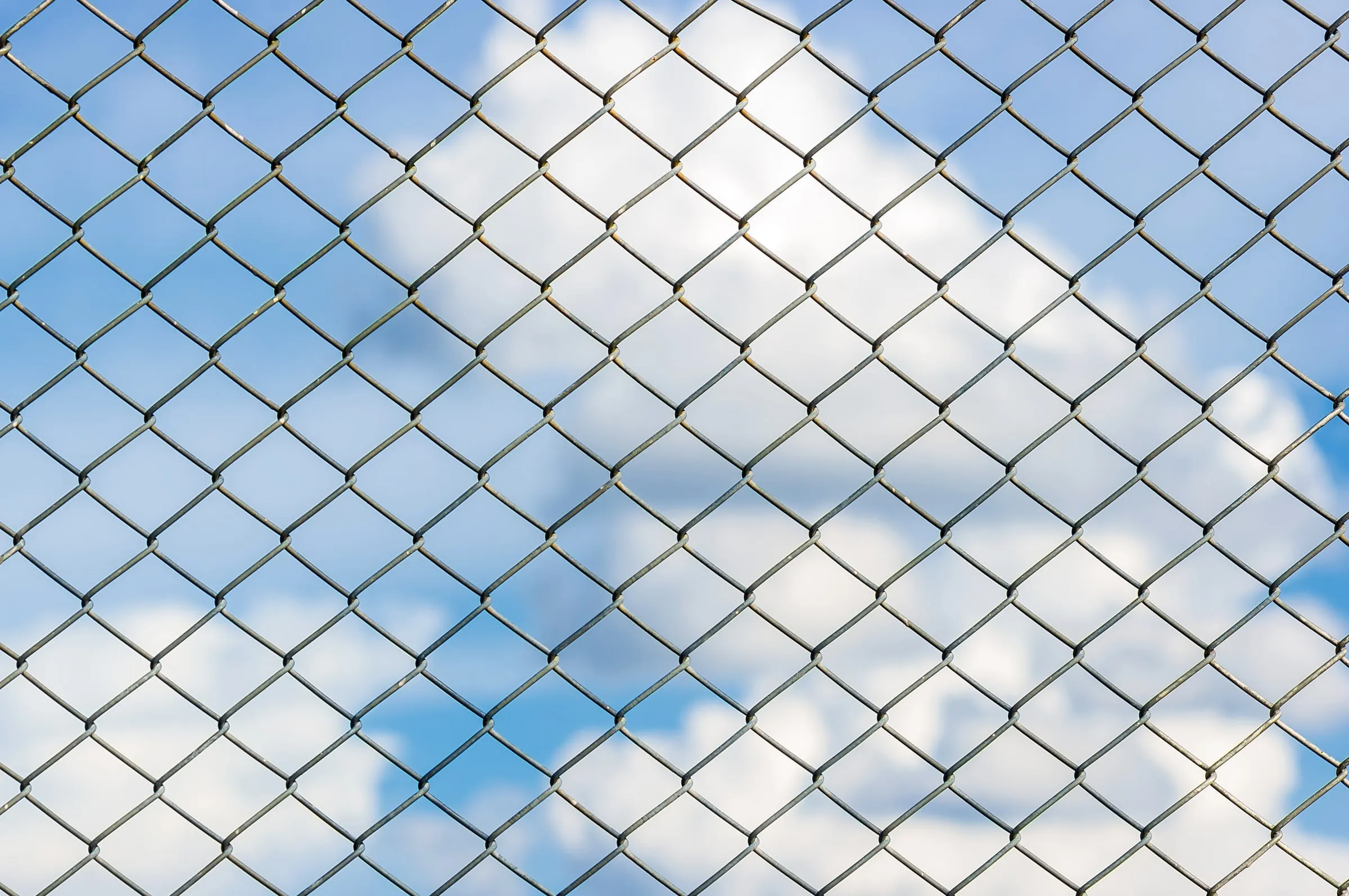 Chain-link fence seen close up against a blue sky with clouds
