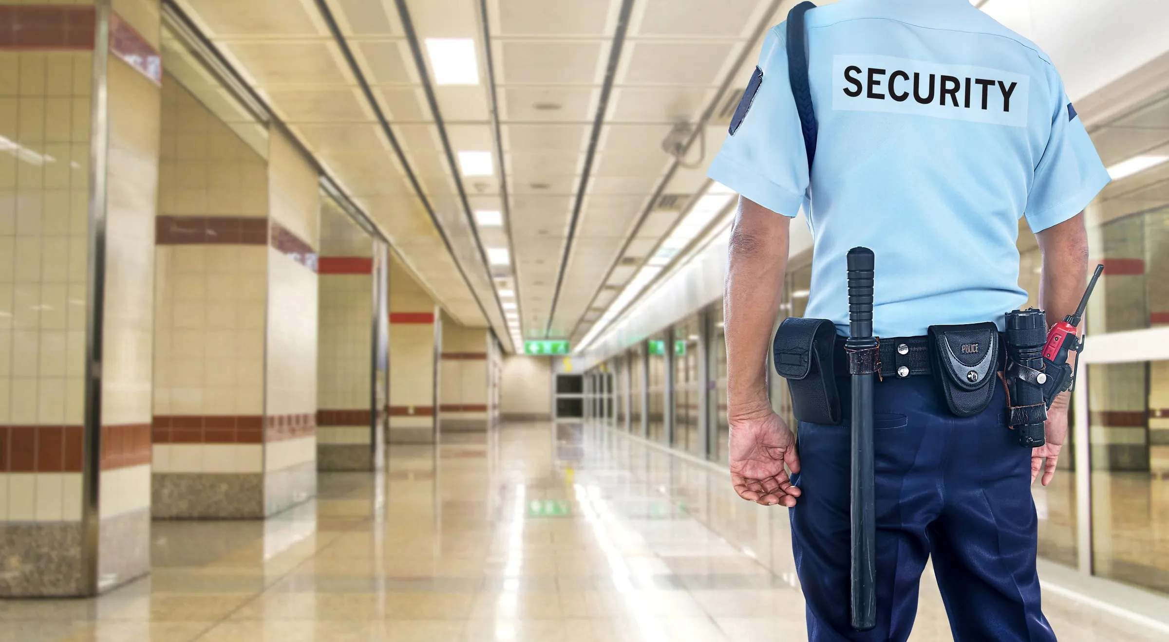 Security officer in a light blue shirt with a duty belt standing in a long tiled corridor