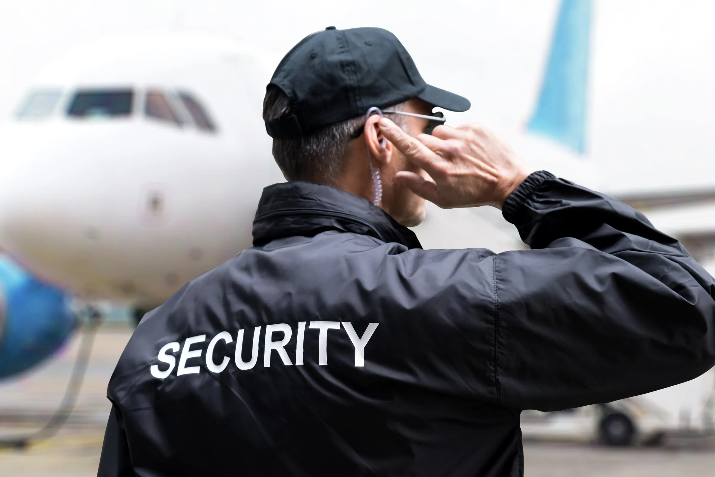 Security officer in a black SECURITY jacket adjusting an earpiece with an aircraft behind