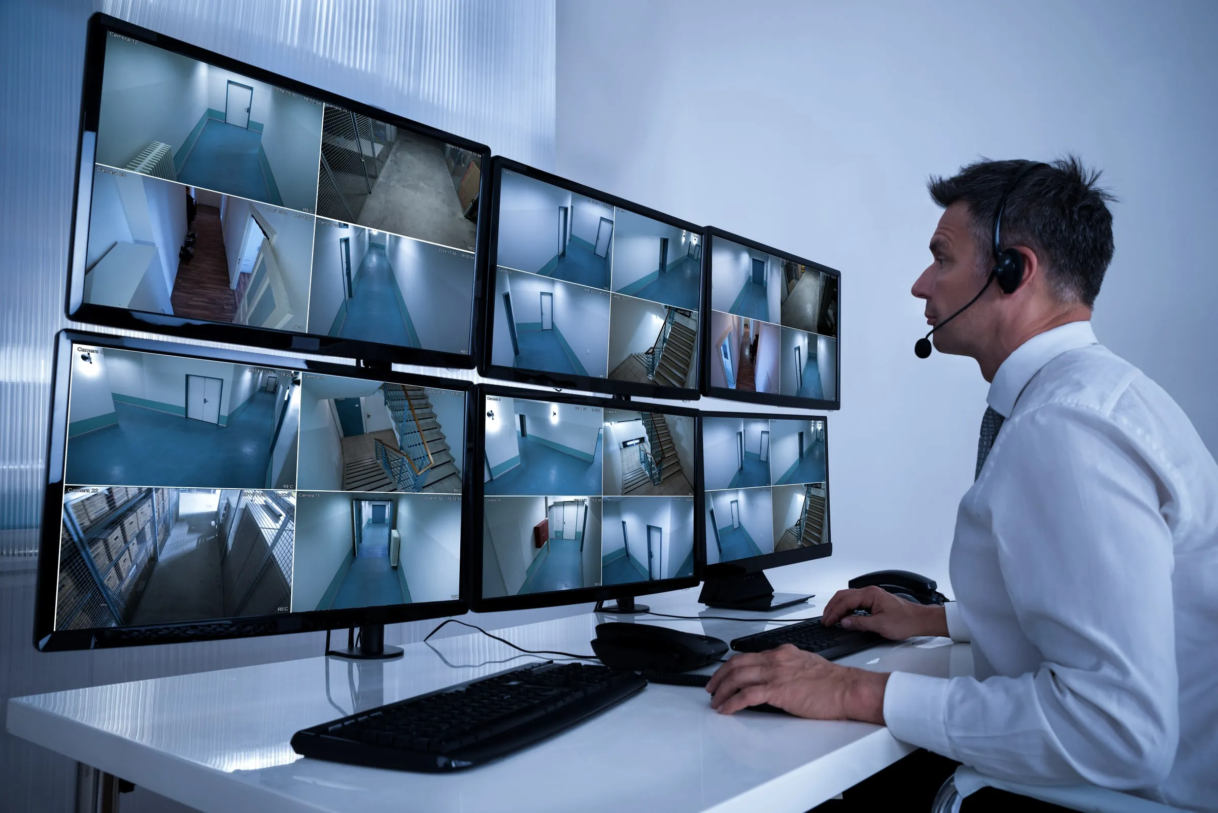 Man with a headset at a desk watching a grid of camera monitors