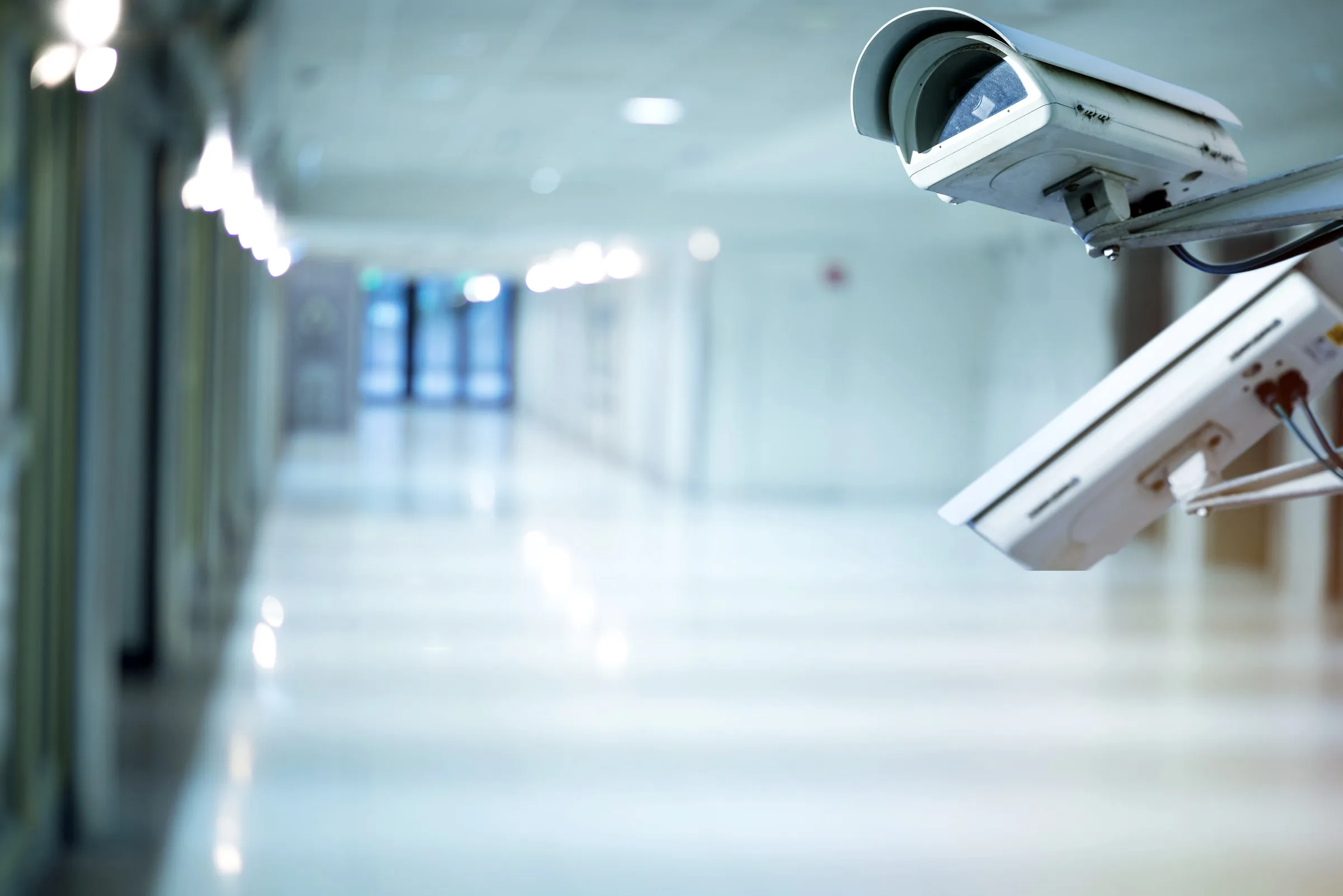 White box-style security camera mounted high on a wall in a bright, empty corridor with glass doors at the far end