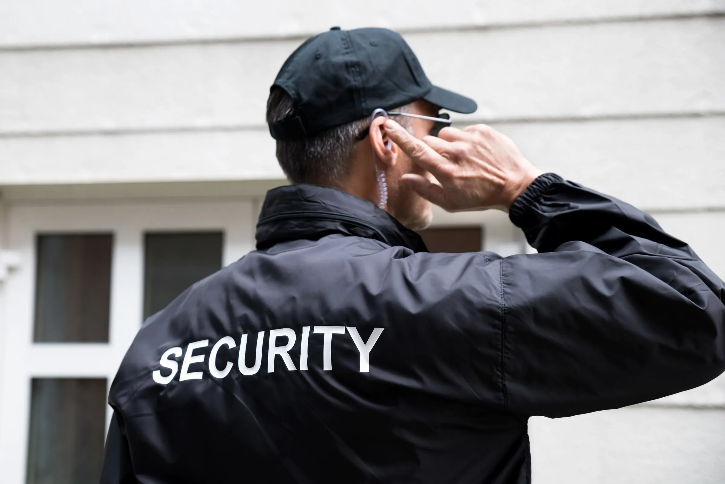 Security officer seen from behind in a black cap and SECURITY jacket pressing his earpiece outside a white building