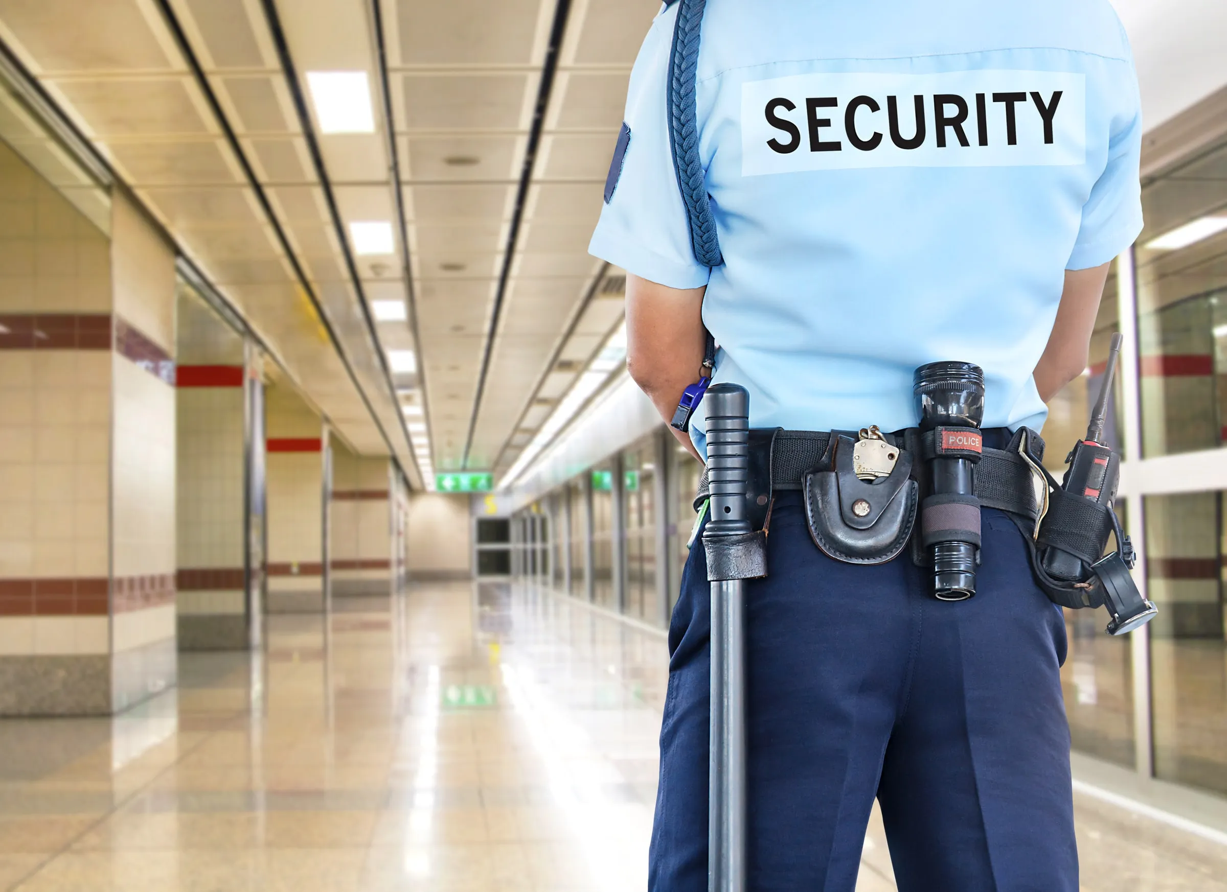 A security officer seen from behind wears a light blue shirt marked security with a duty belt, flashlight and radio in a corridor