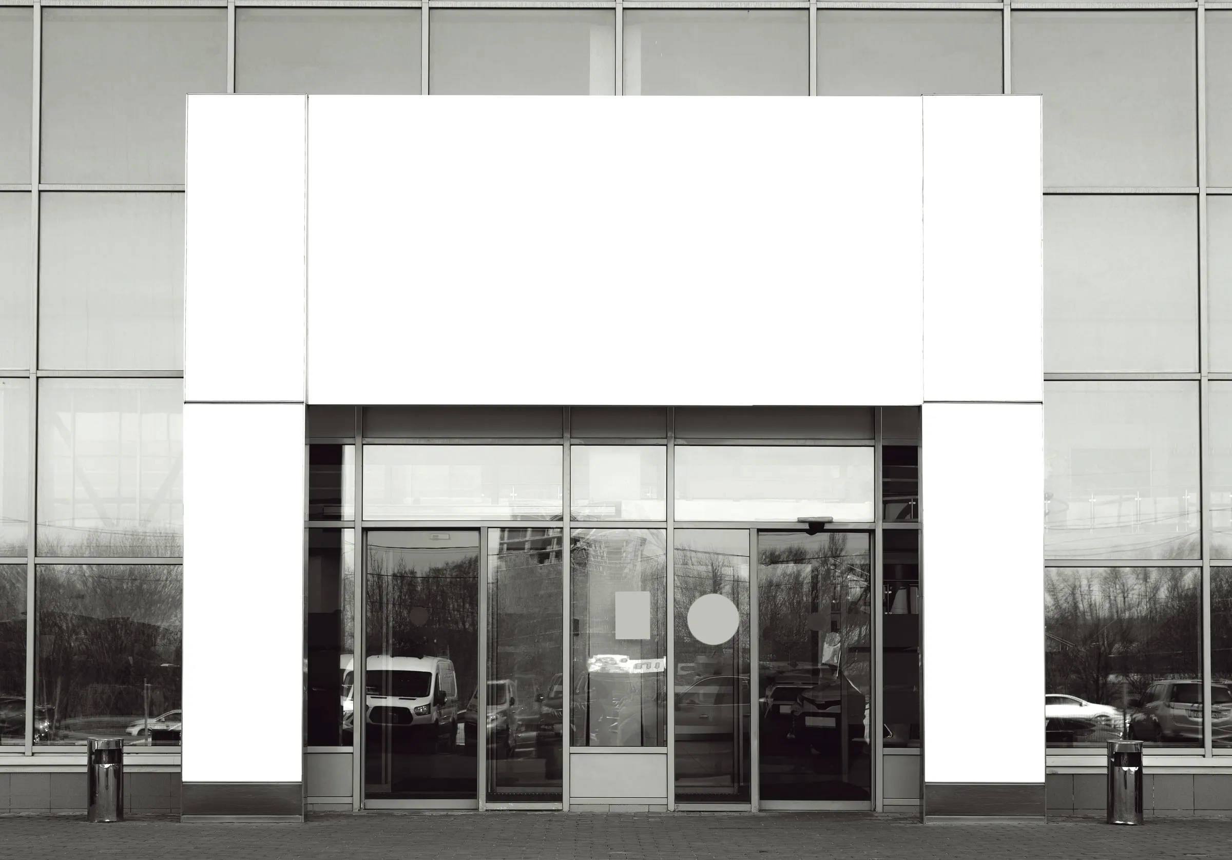 A blank white illuminated sign panel above the glass sliding entrance doors of a big-box store, shown in black and white
