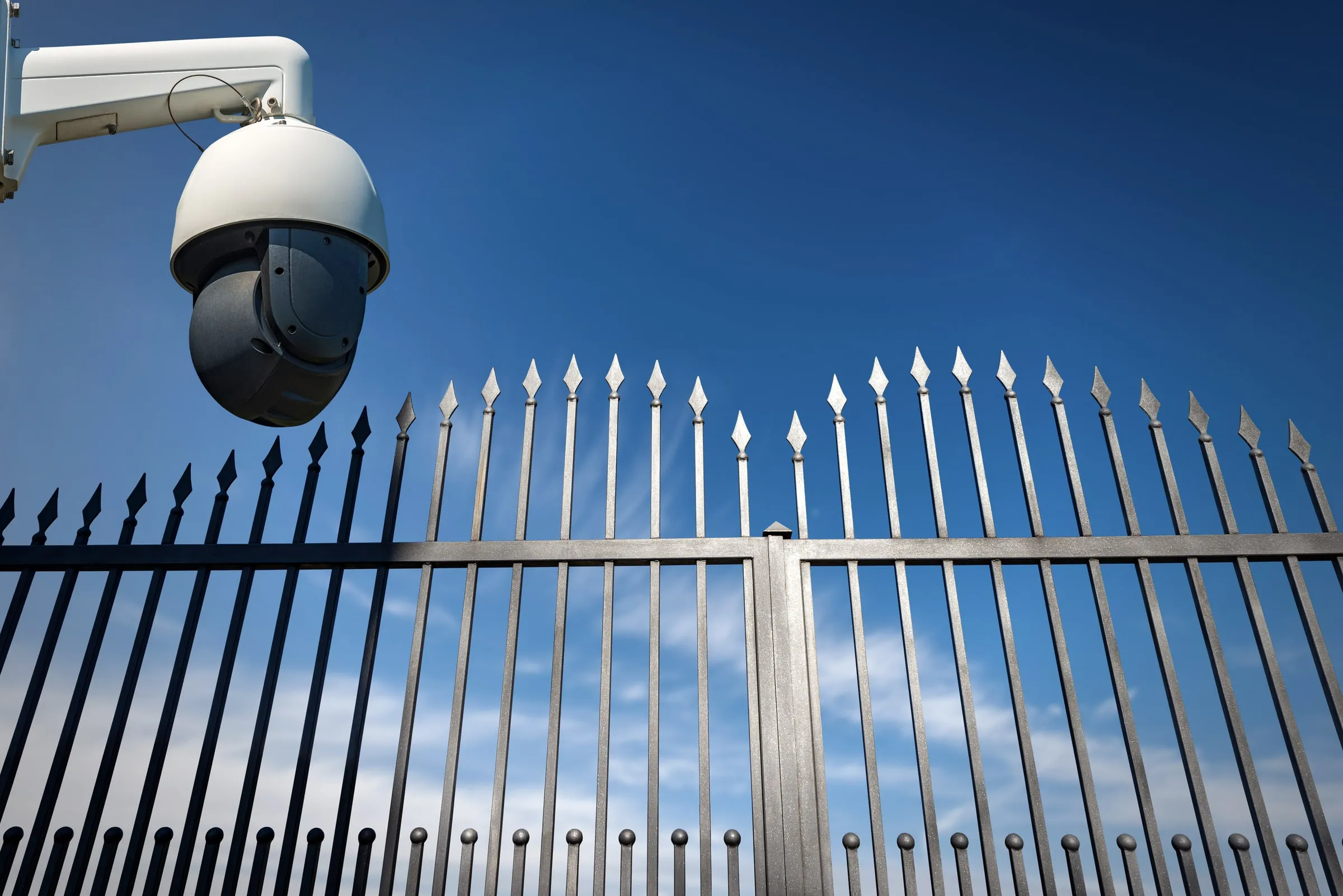 A security camera dome mounts on an exterior wrought-iron fence against a clear blue sky