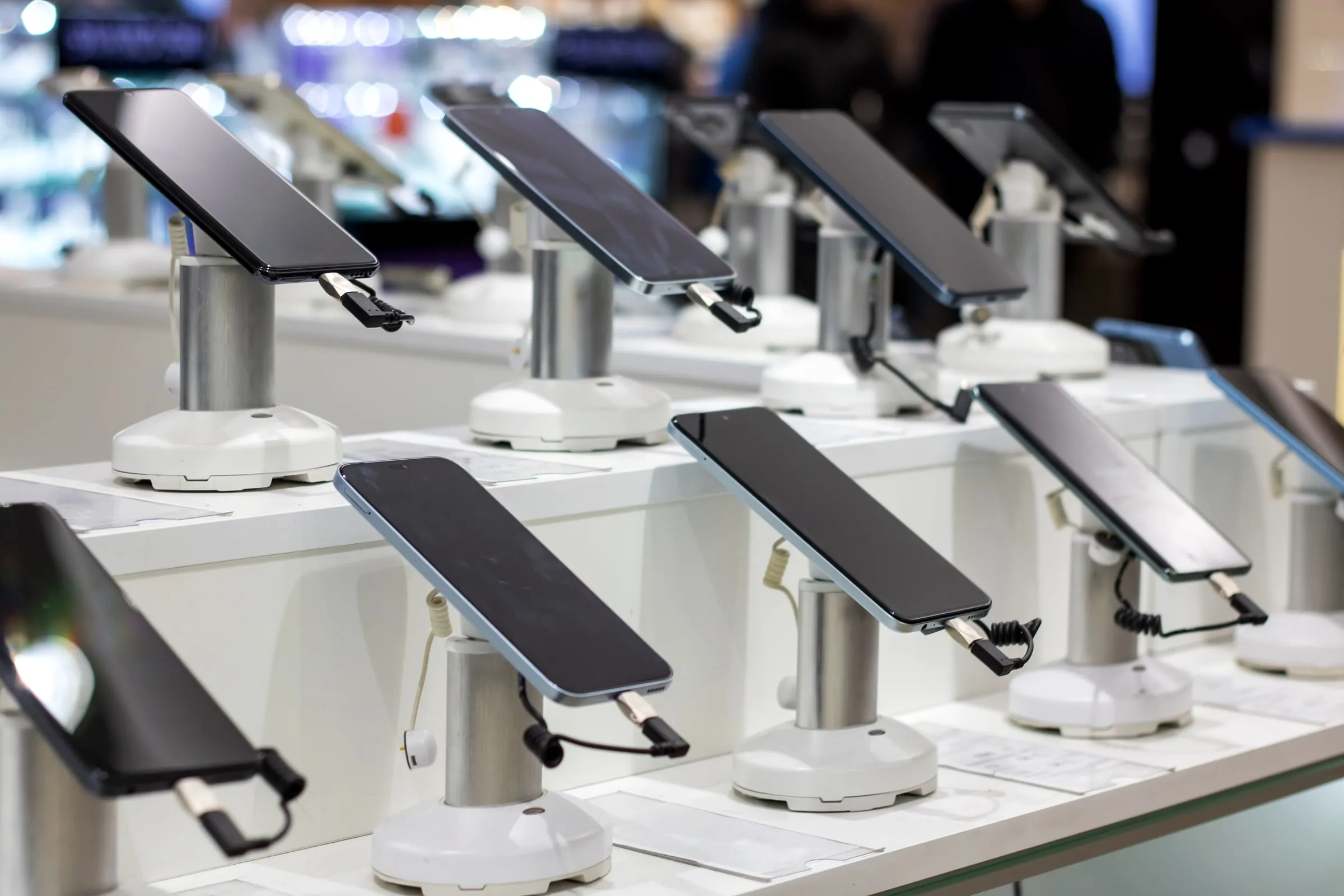 A row of smartphones mounted on anti-theft security stands with charging cables, displayed on a store counter