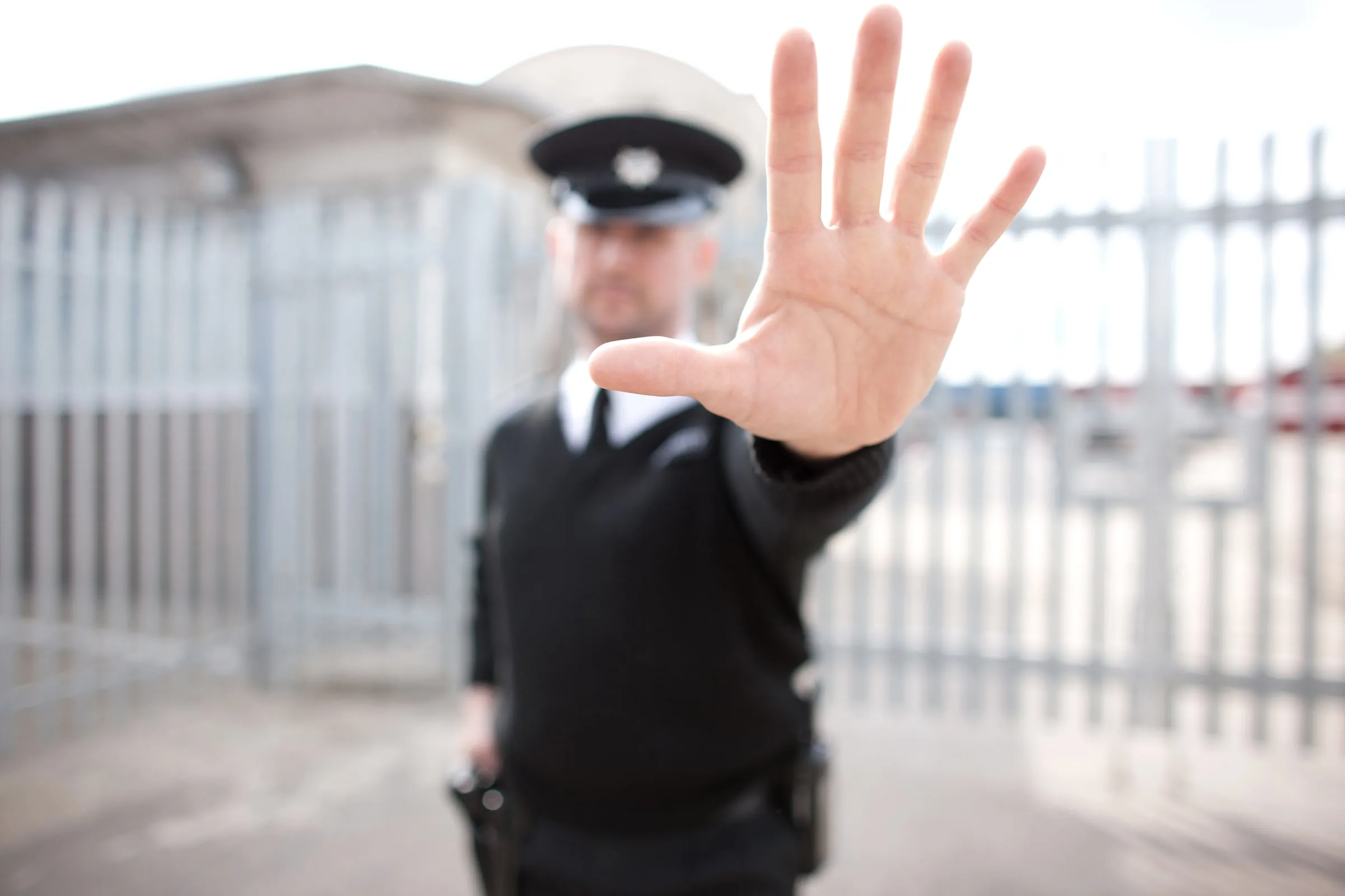 Uniformed security officer in a peaked cap raising an open palm in a stop gesture in front of a metal gate
