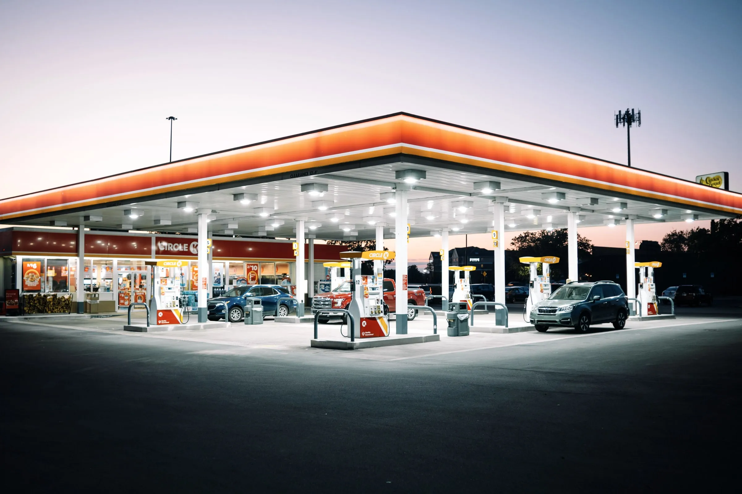 Lit gas station canopy over fuel pumps at dusk, with cars refueling and a convenience store front behind them