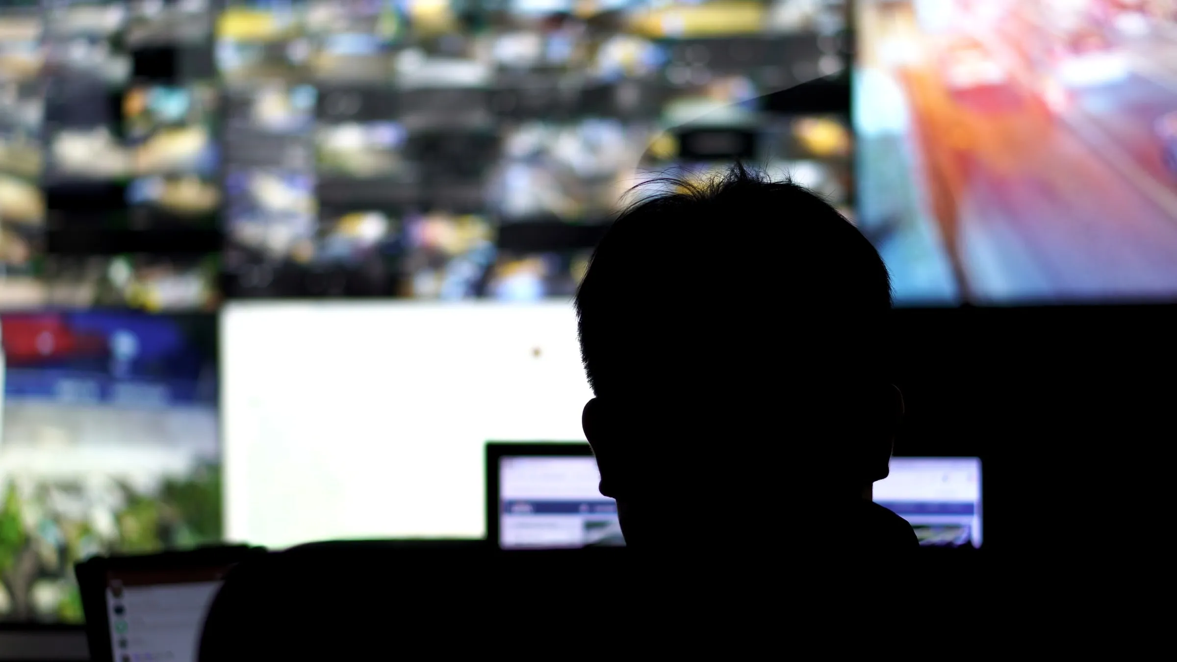 Silhouette of a person seated in a dark room facing a wall of blurred camera monitors